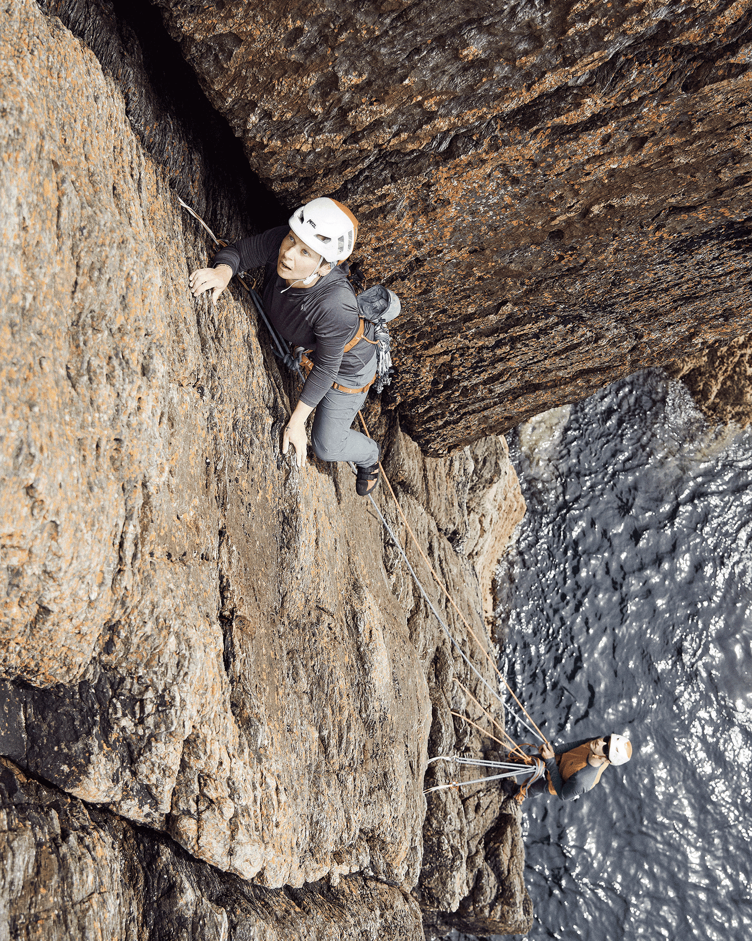 Two climbers scale a rugged sea cliff, roped together, wearing helmets and harnesses with the ocean visible below.