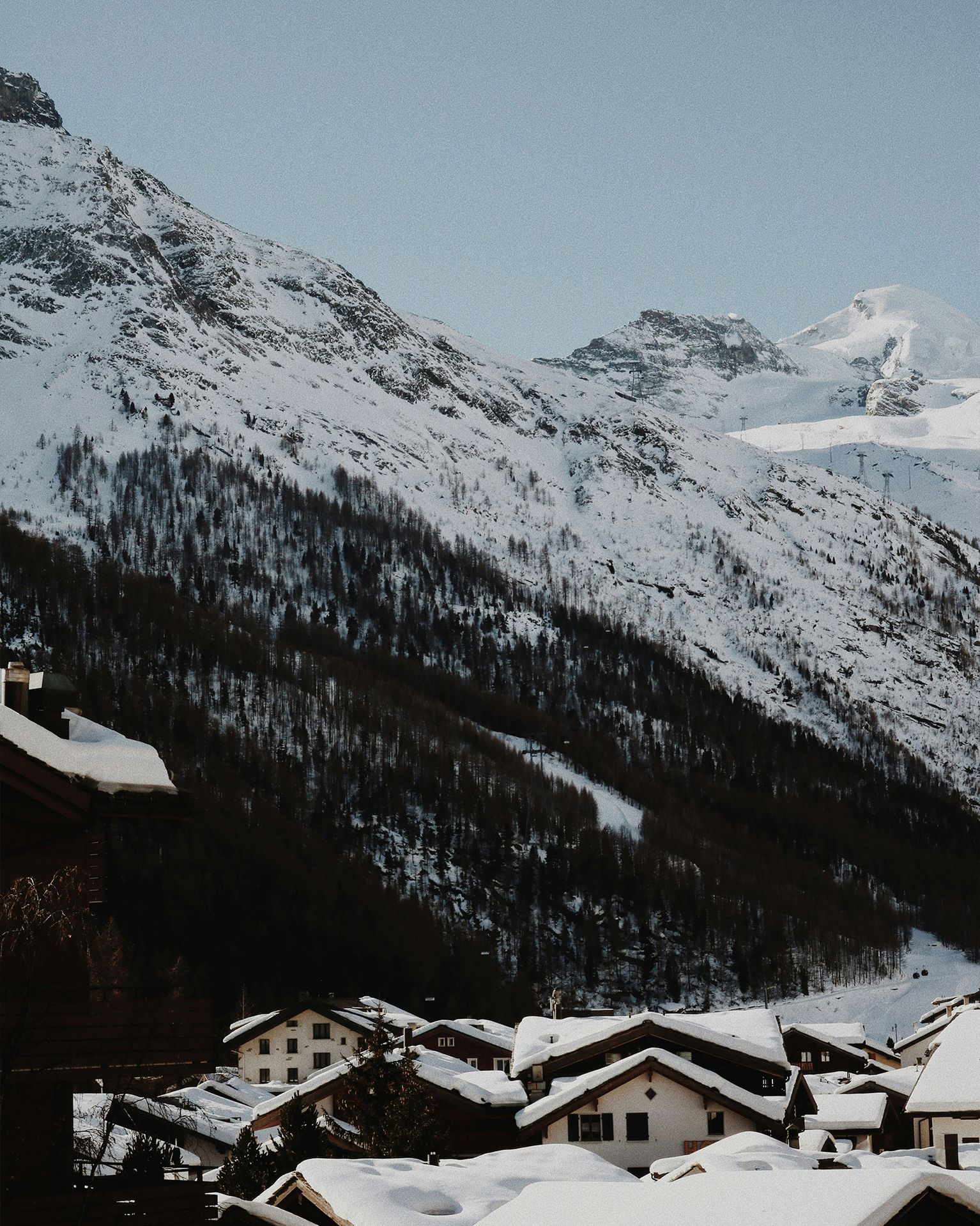 Snow-covered mountain landscape with a cluster of chalet-style houses in the foreground, under a clear blue sky.