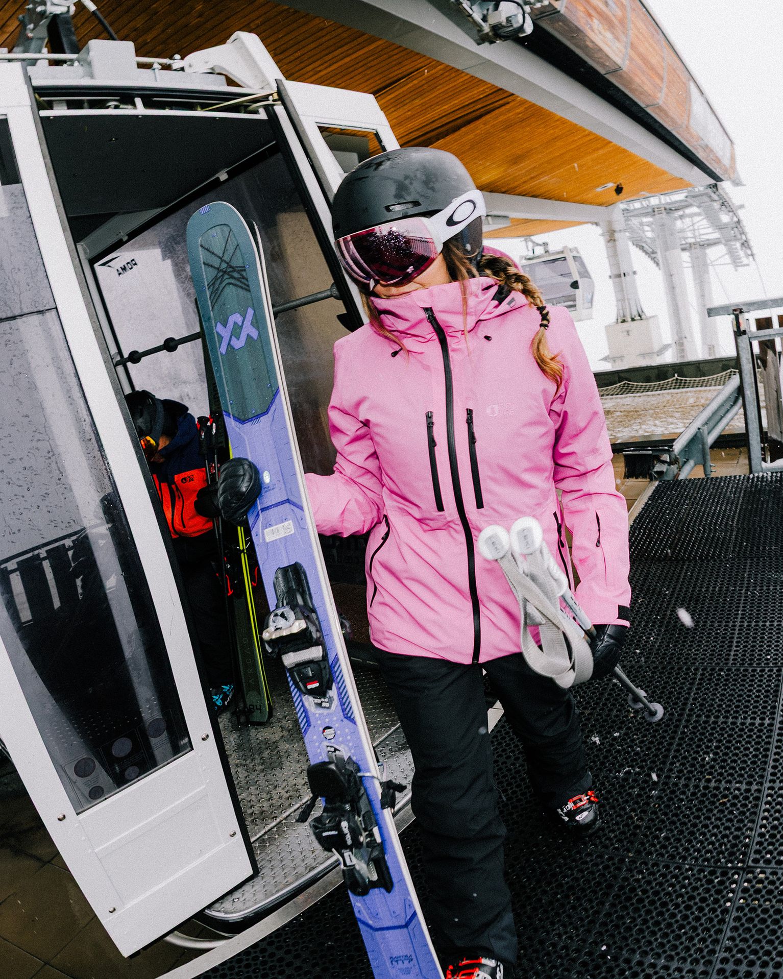 A person in a pink jacket and helmet exits a ski lift, holding skis and poles, ready for mountain skiing.