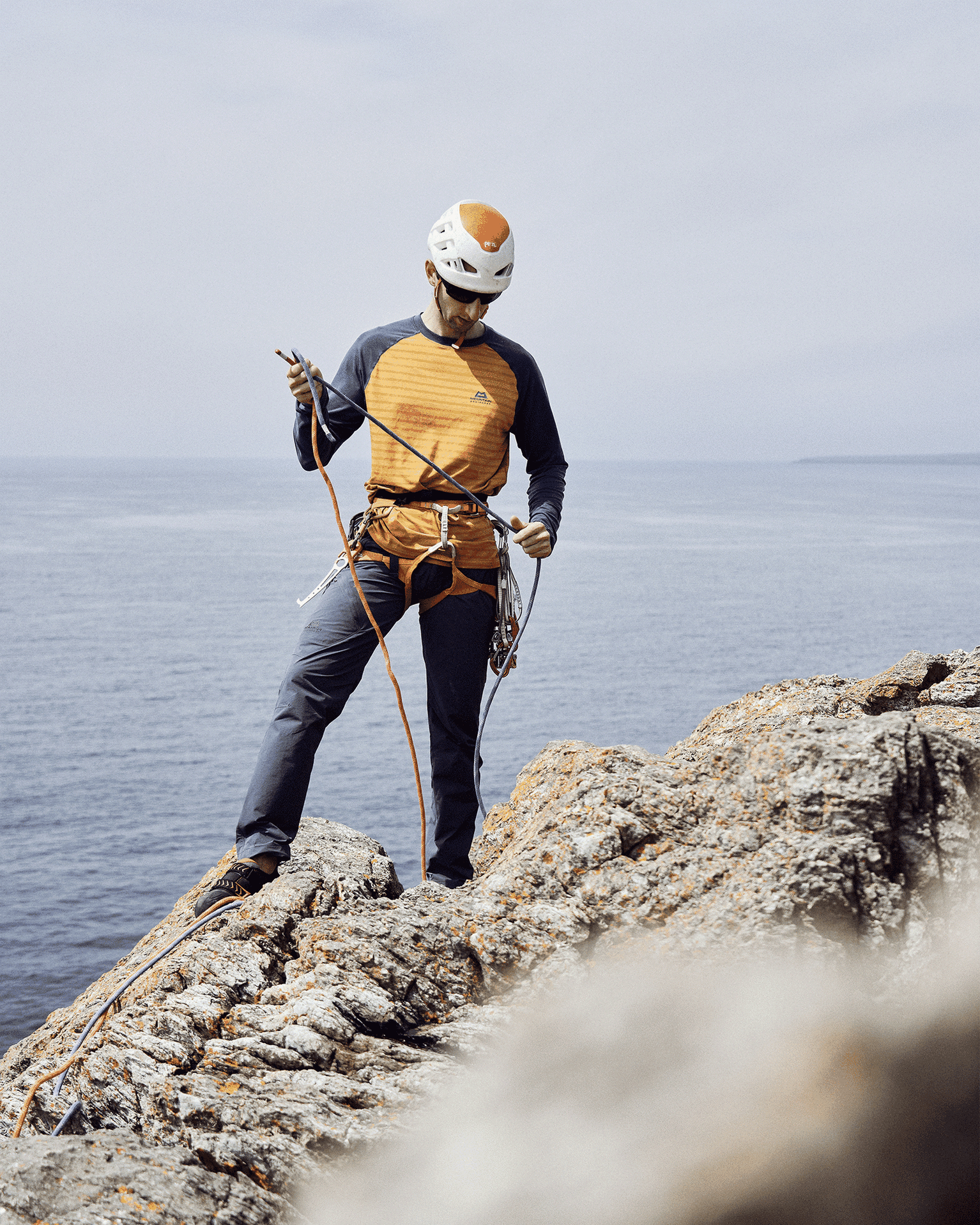 Climber wearing a helmet and harness stands on a rocky cliff, adjusting ropes, with the ocean in the background.