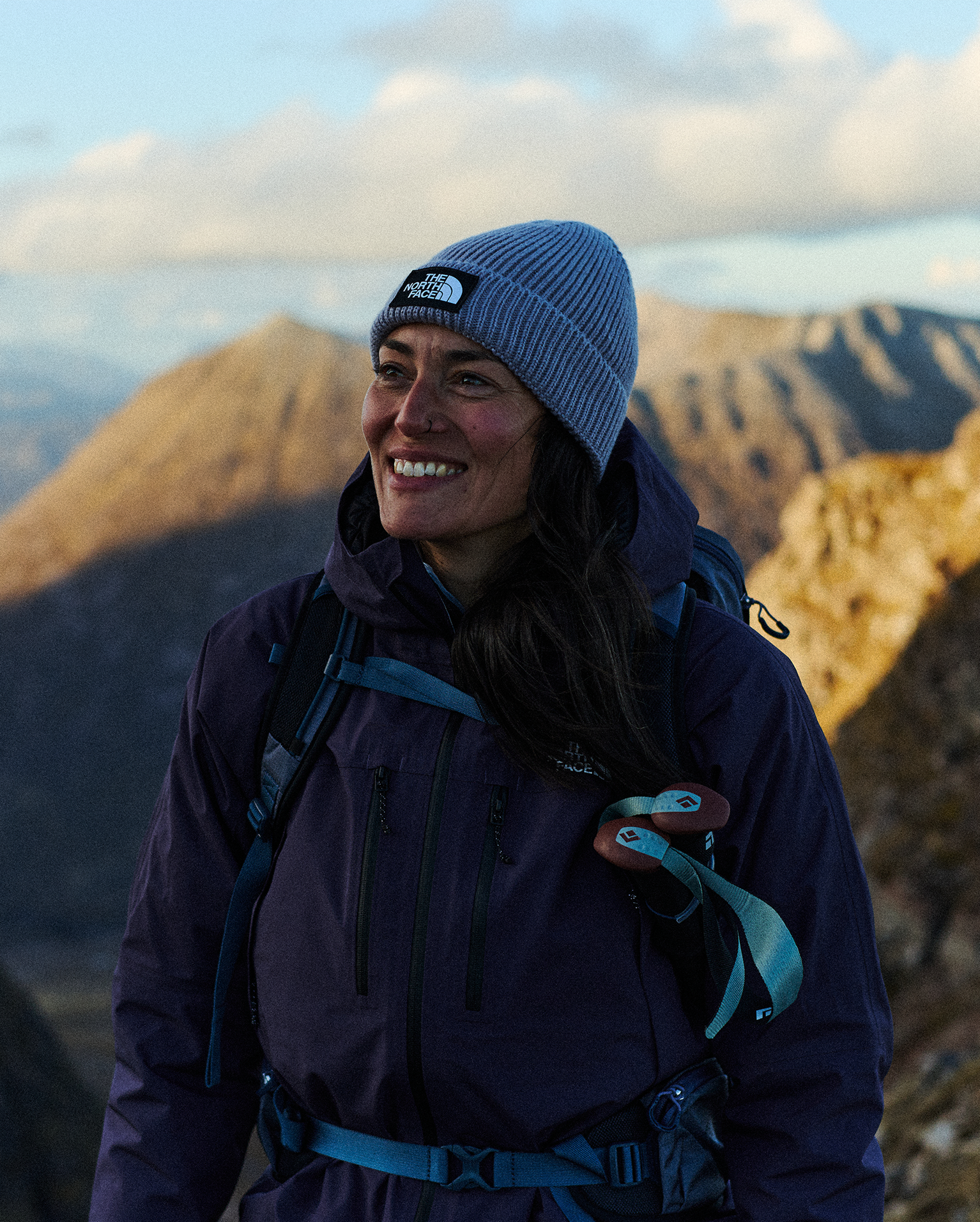 Smiling woman in a beanie and outdoor jacket stands on a mountain trail, rugged peaks and soft clouds in the background.