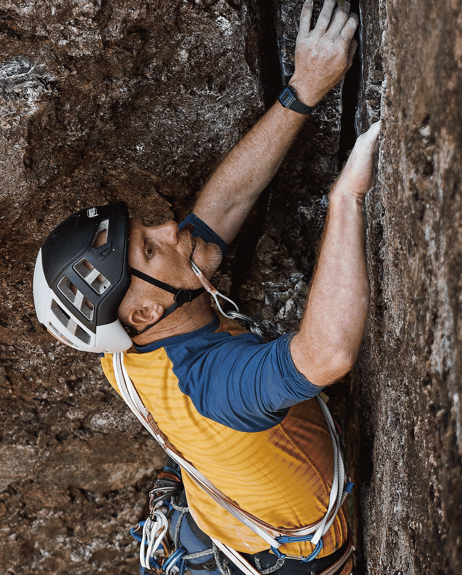 Rock climber with helmet and gear scaling a rocky surface, gripping tightly and wearing a yellow and blue top.