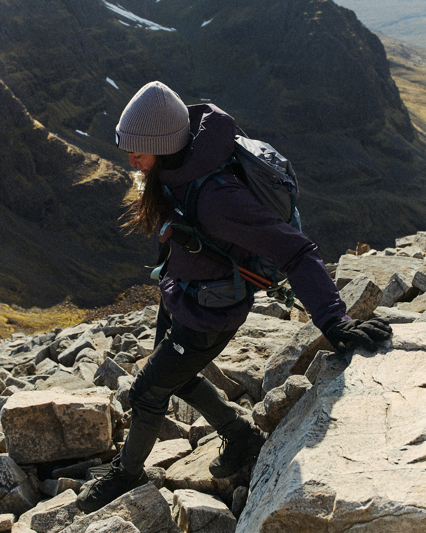 A hiker with a backpack and beanie climbs over rocky terrain in mountainous landscape, showcasing determination and rugged outdoor skills.