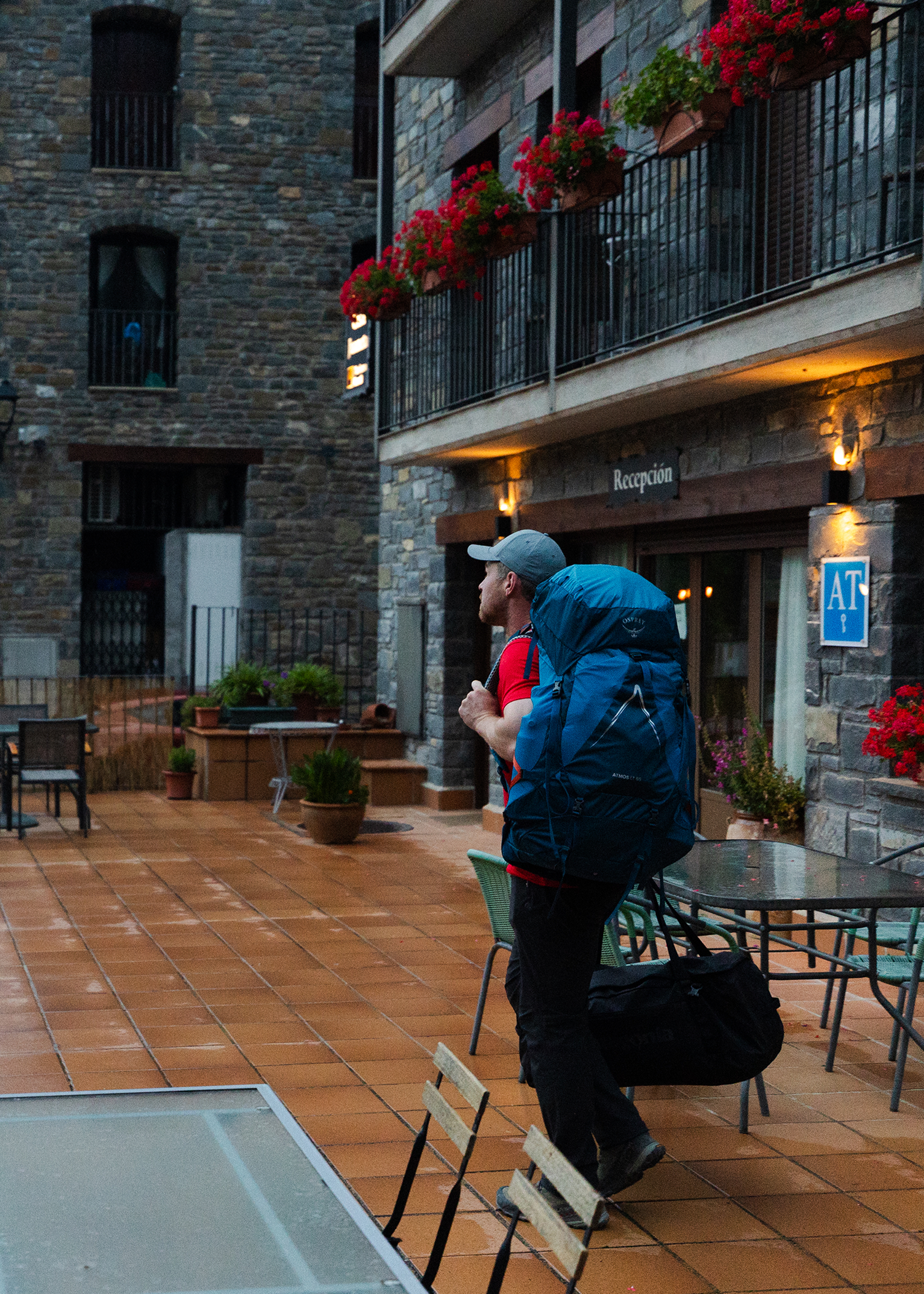 A person with a large blue backpack stands on a tiled patio outside a stone building with red flowers and a sign reading "Reception."