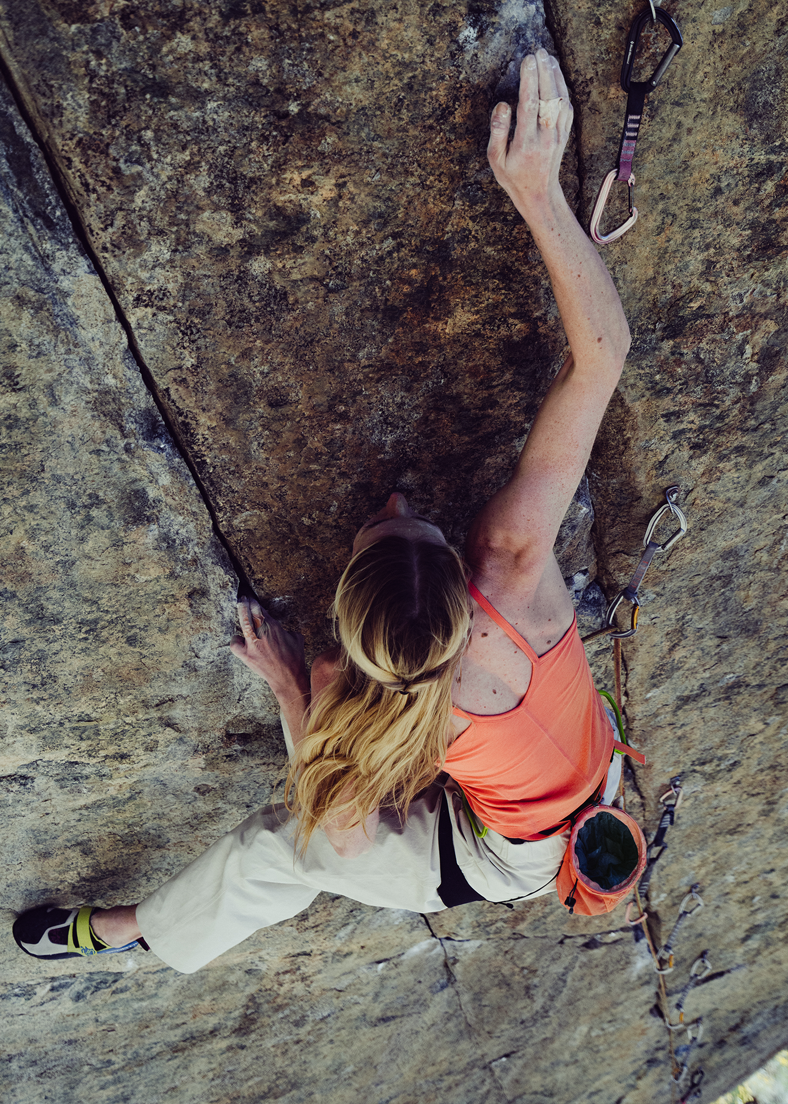 A climber wearing a coral tank top and white trousers ascends a steep rock face, demonstrating strength and skill.