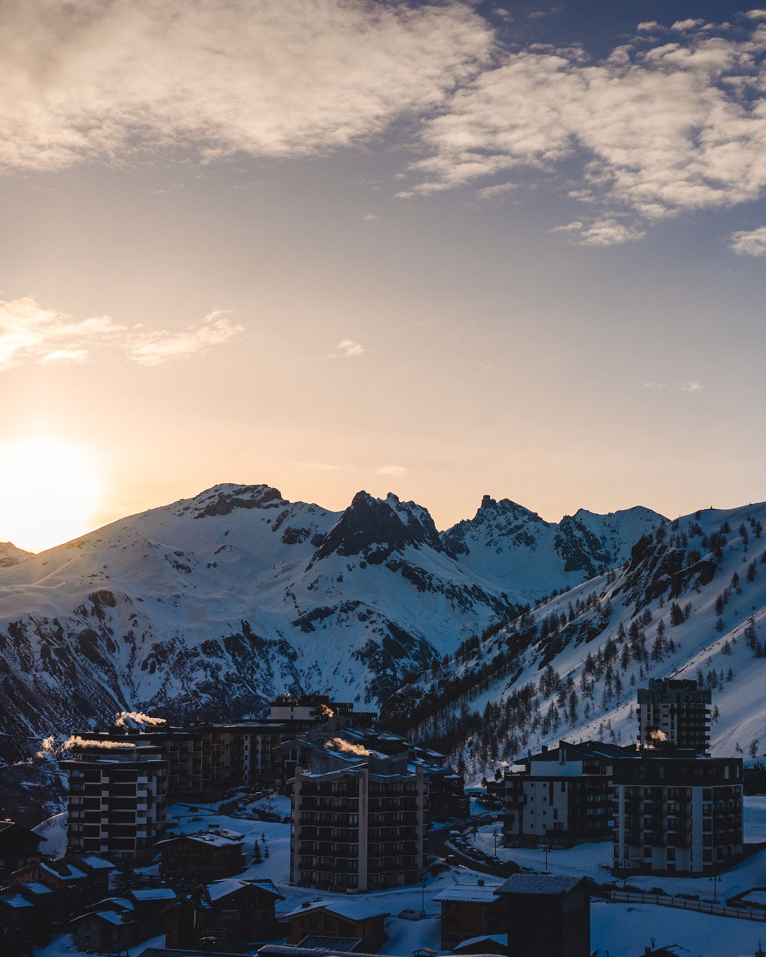 Sun setting behind snow-covered mountains, casting a warm glow over a town with tall buildings in the foreground.