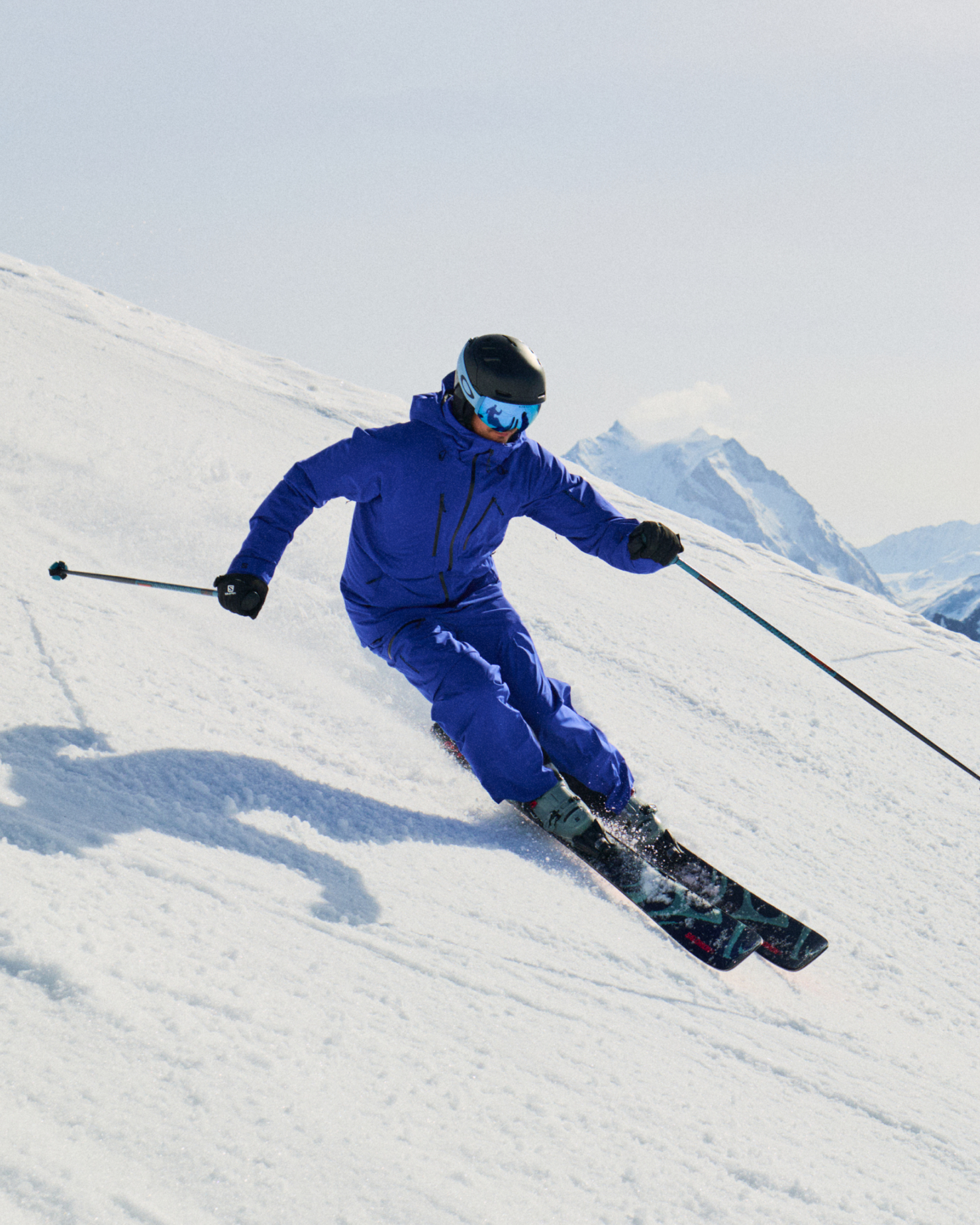Skier carving down a snowy mountain slope, dressed in a blue jacket and trousers, wearing a helmet and goggles, with snowy peaks in the background.