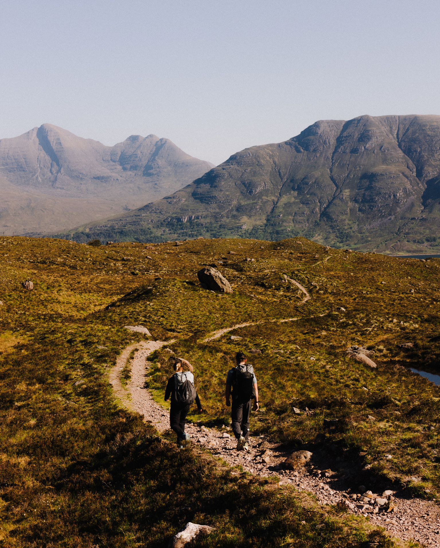 Two hikers walking on a winding trail, surrounded by rugged mountains under a clear blue sky.