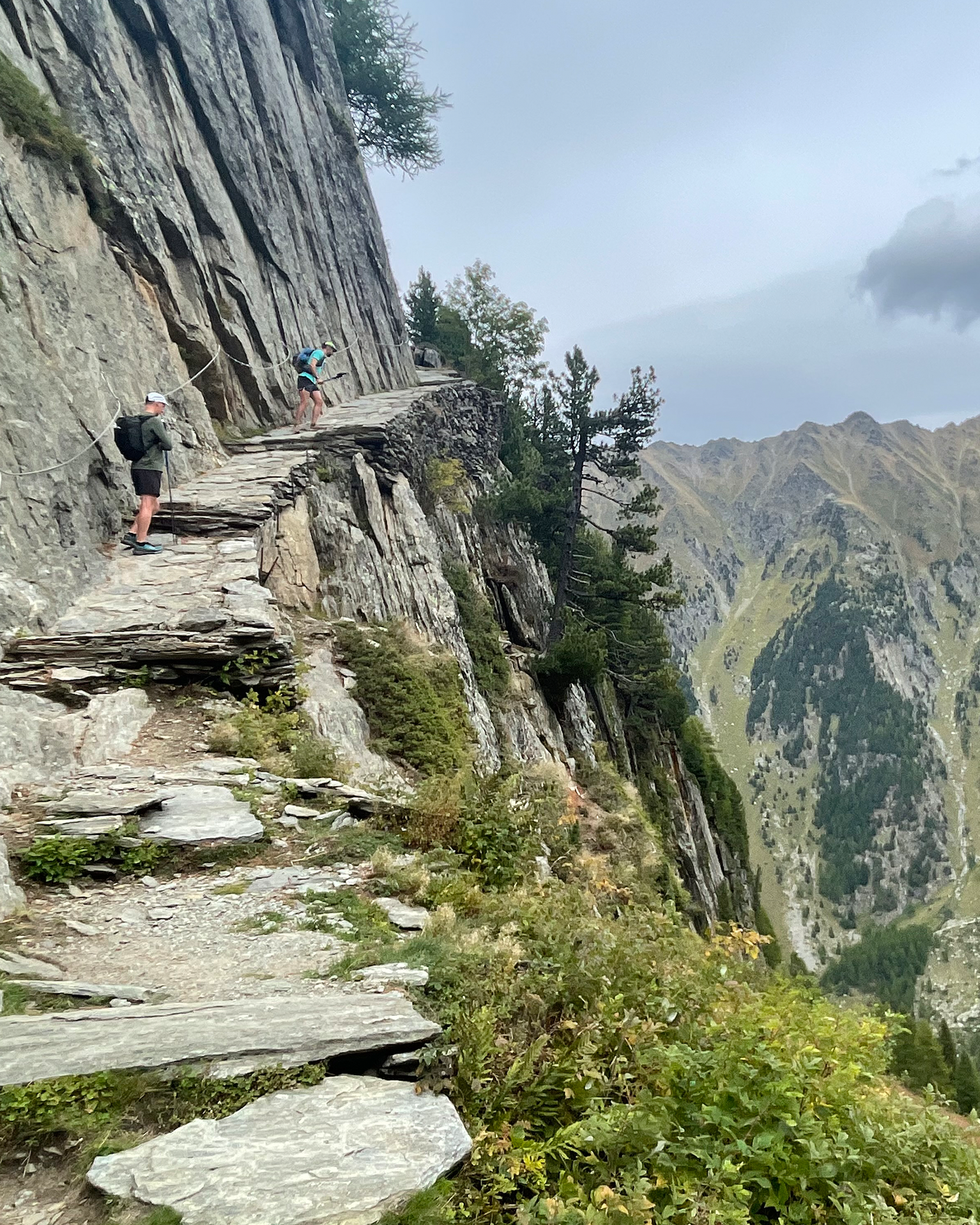 Hikers navigate a narrow, rocky mountain path with steep cliffs and distant peaks under a cloudy sky.