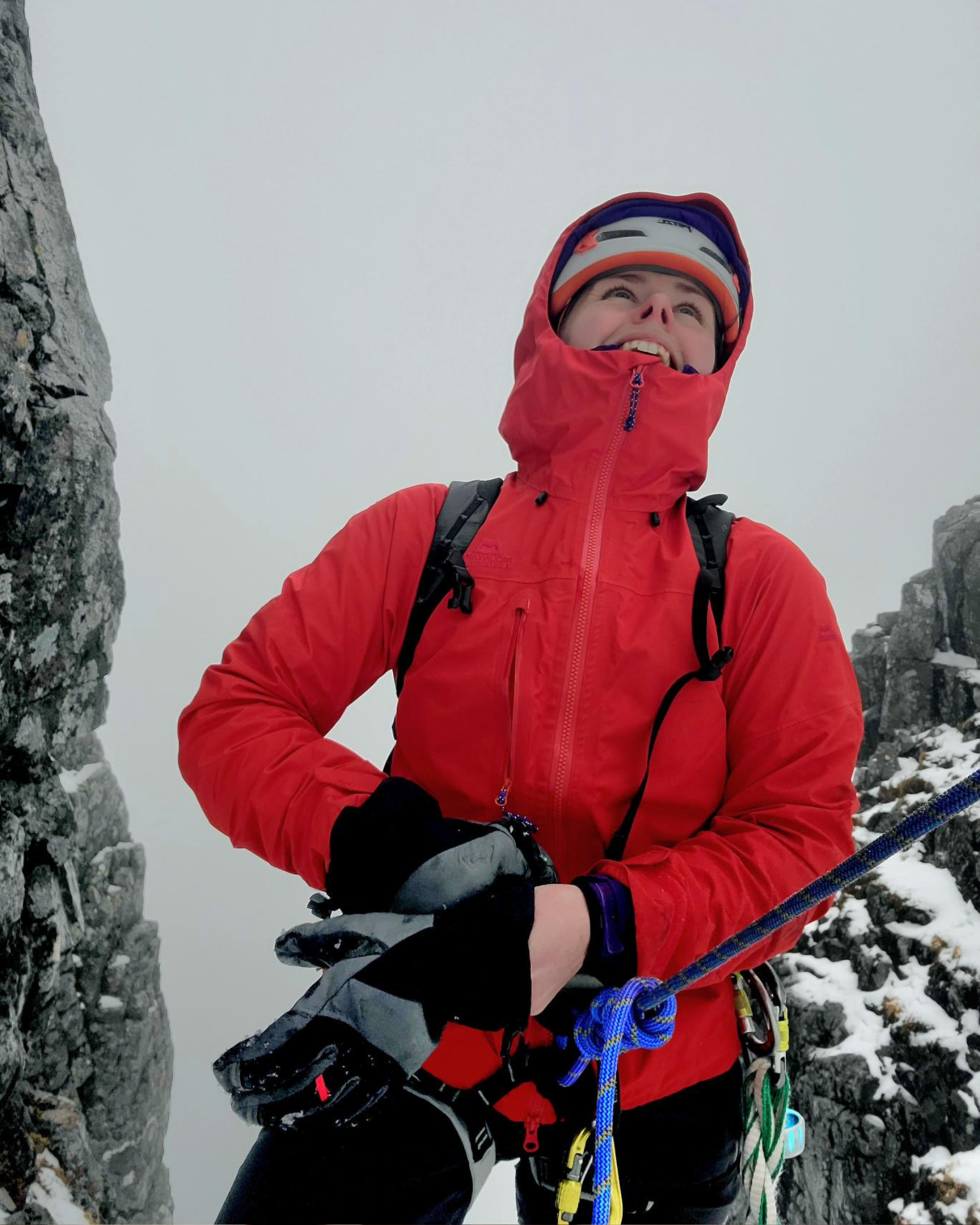 A climber in a red jacket and helmet smiles while holding climbing gear on a snowy mountain.
