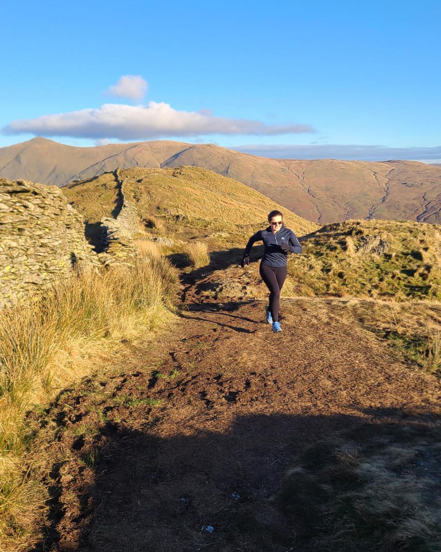 Person running on a scenic mountain trail under a clear blue sky, surrounded by grassy hills and distant peaks.