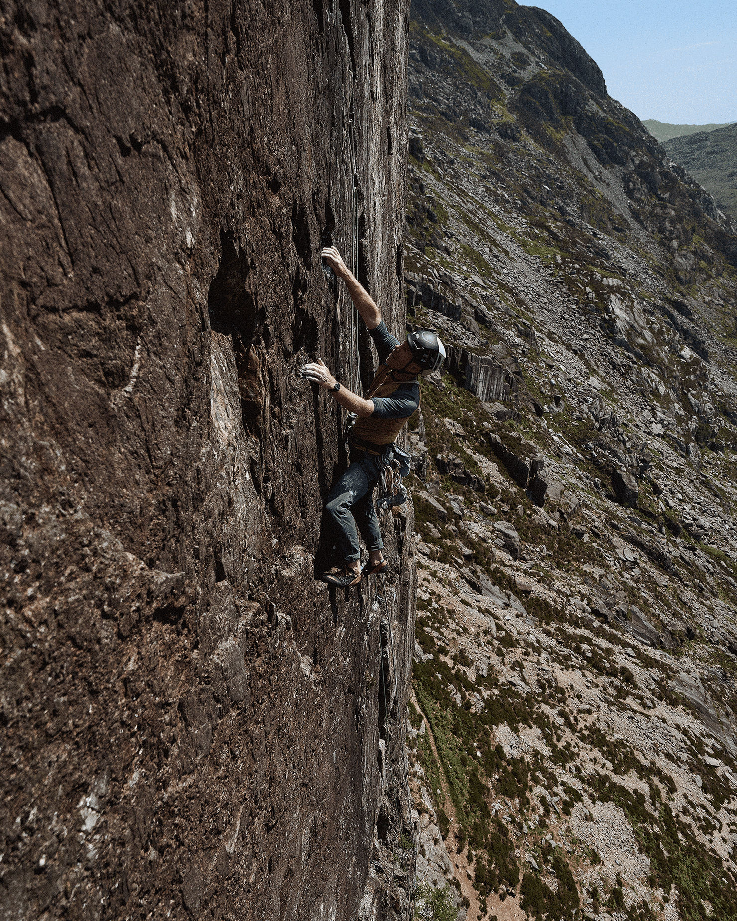 Climber scaling a steep rock face with rugged mountain terrain in the background, wearing a helmet and climbing gear.