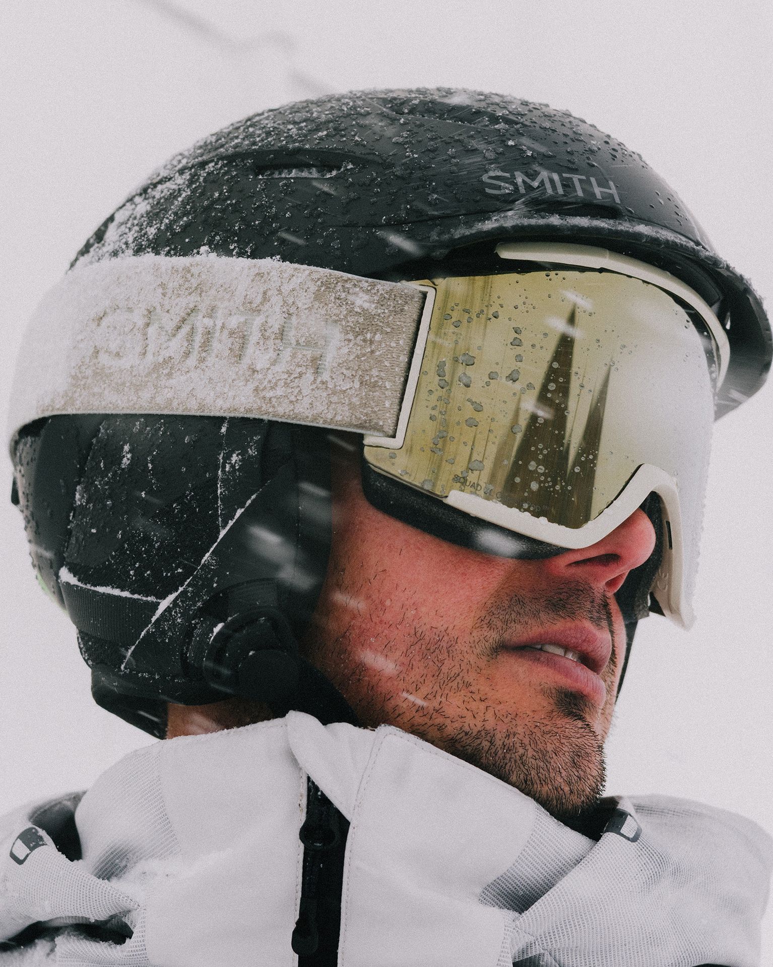 Person in a snow-covered helmet and goggles, wearing a white jacket, looking sideways in a snowy environment.