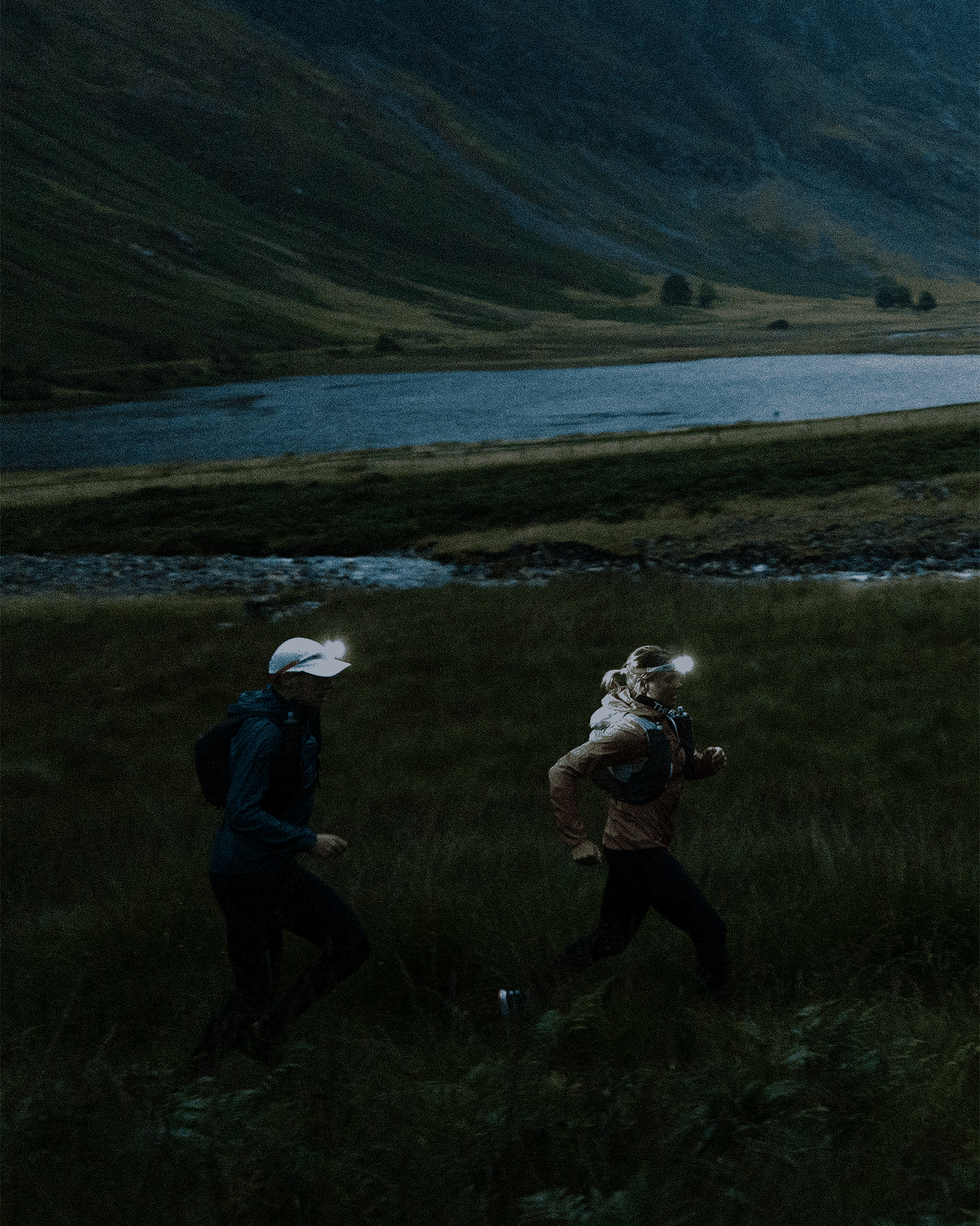 Two people trail running at dusk, wearing head torches and outdoor gear, with a mountain and lake in the background.