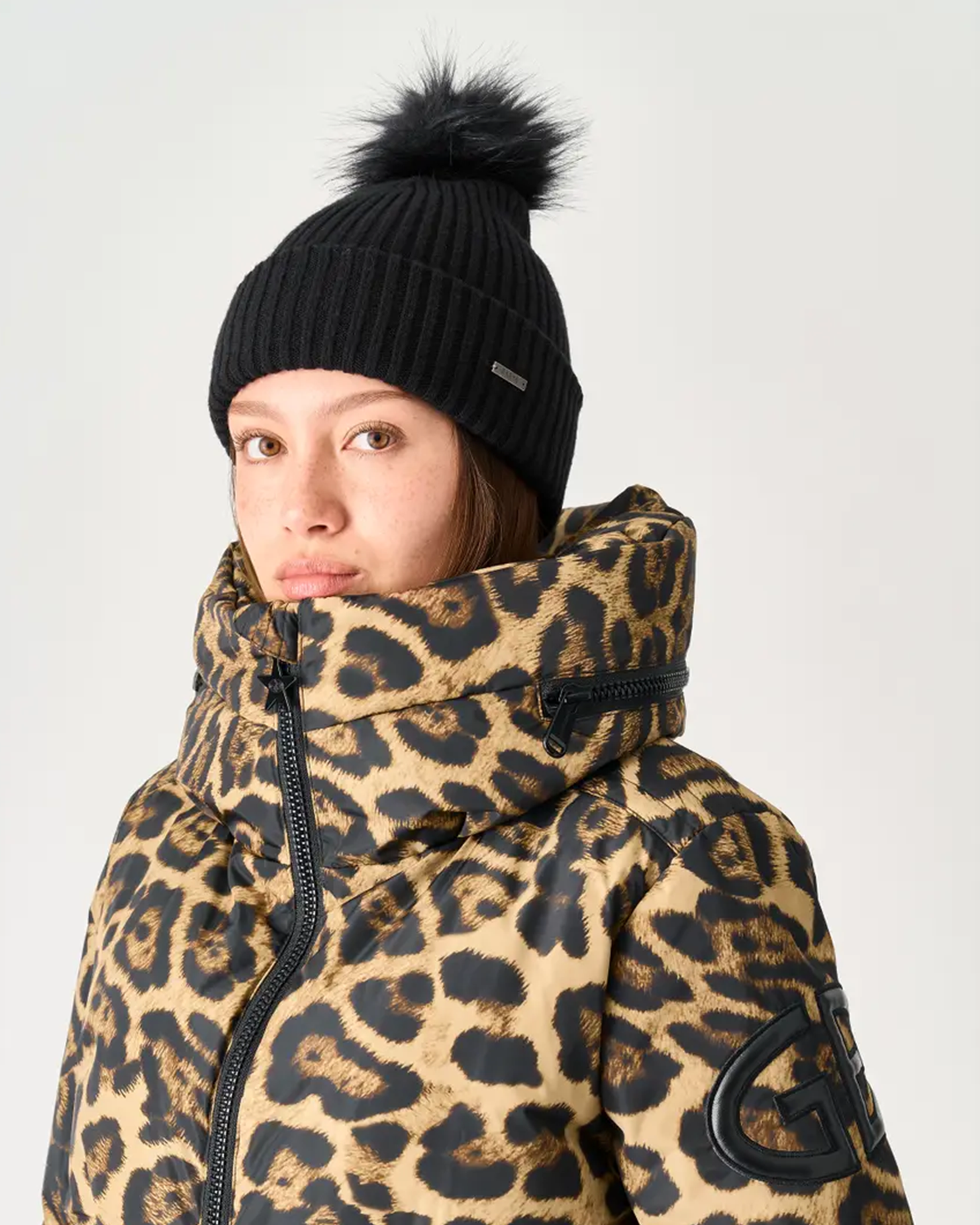 Person wearing insulated leopard-print high-collar jacket and black ribbed beanie with faux-fur pompom against a plain studio background.