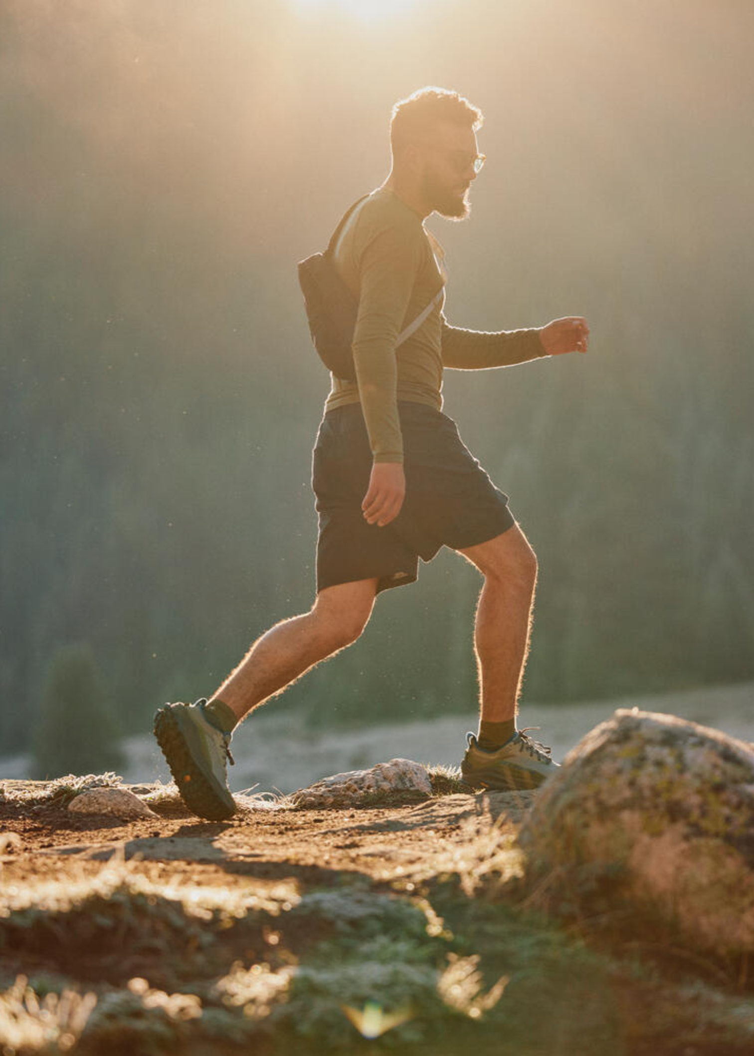 Man in outdoor gear hiking on a rocky trail at sunrise, surrounded by trees and warm sunlight.