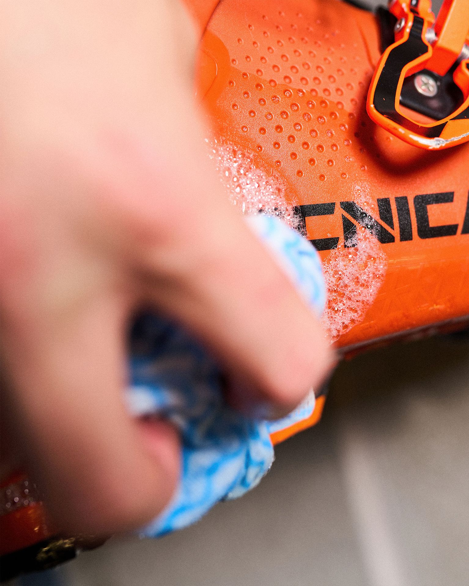 Close-up of a person cleaning an orange ski boot with a blue cloth, showing soap suds and part of the boot's branding.