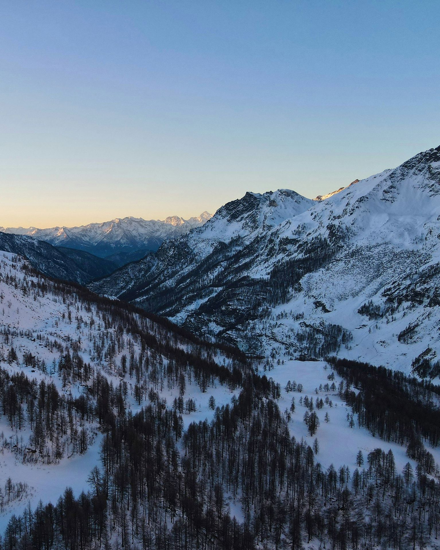 Snow-covered mountain landscape with a forested valley at sunset, featuring distant peaks under a clear sky.