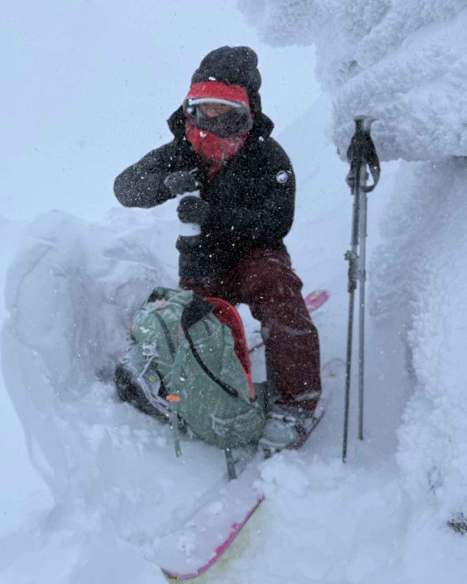 A person in winter gear and goggles rests with a backpack on snowy terrain, holding a drink beside ski poles.
