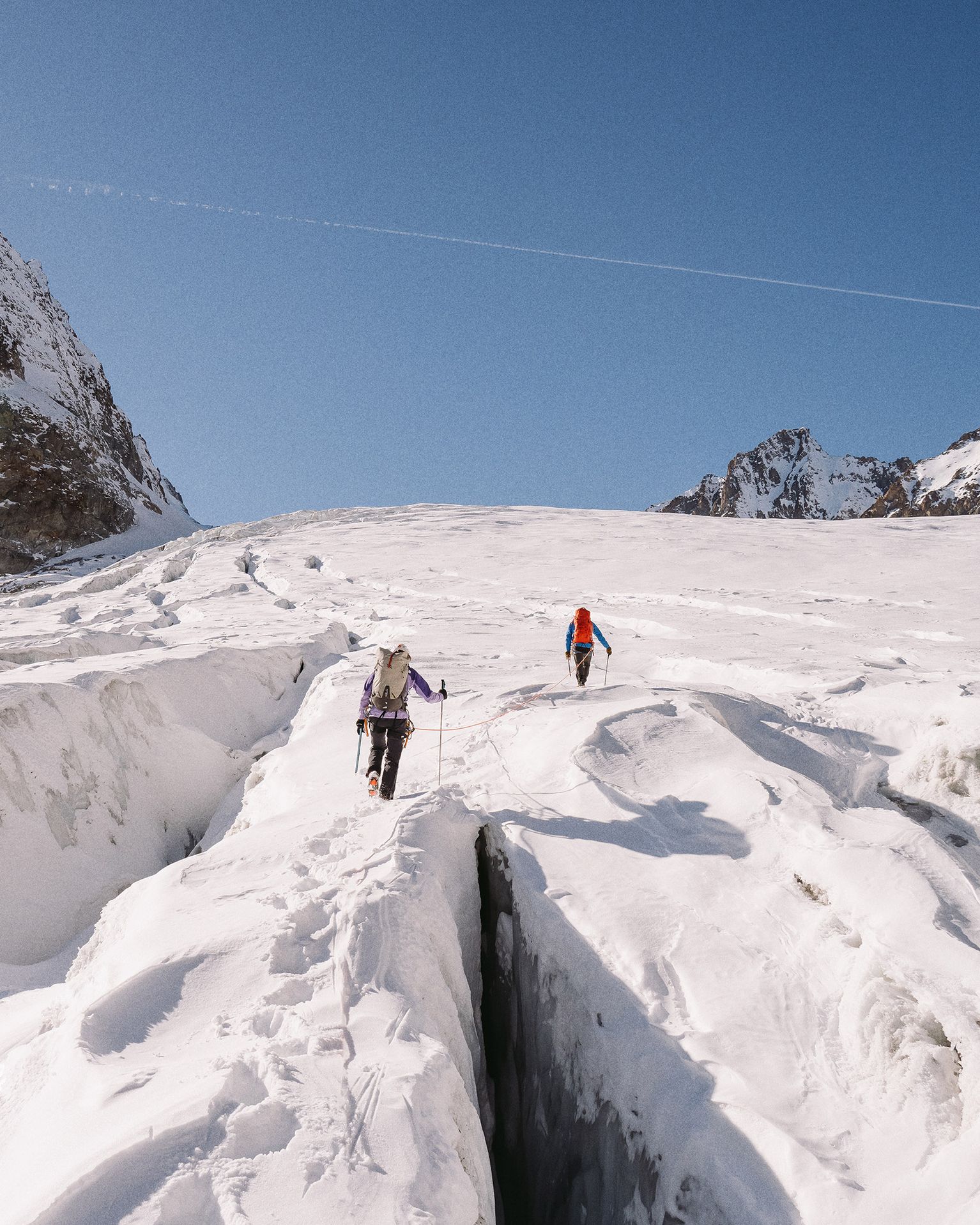 Two climbers with backpacks navigate a snowy mountain landscape, crossing a deep crevasse under a clear blue sky.
