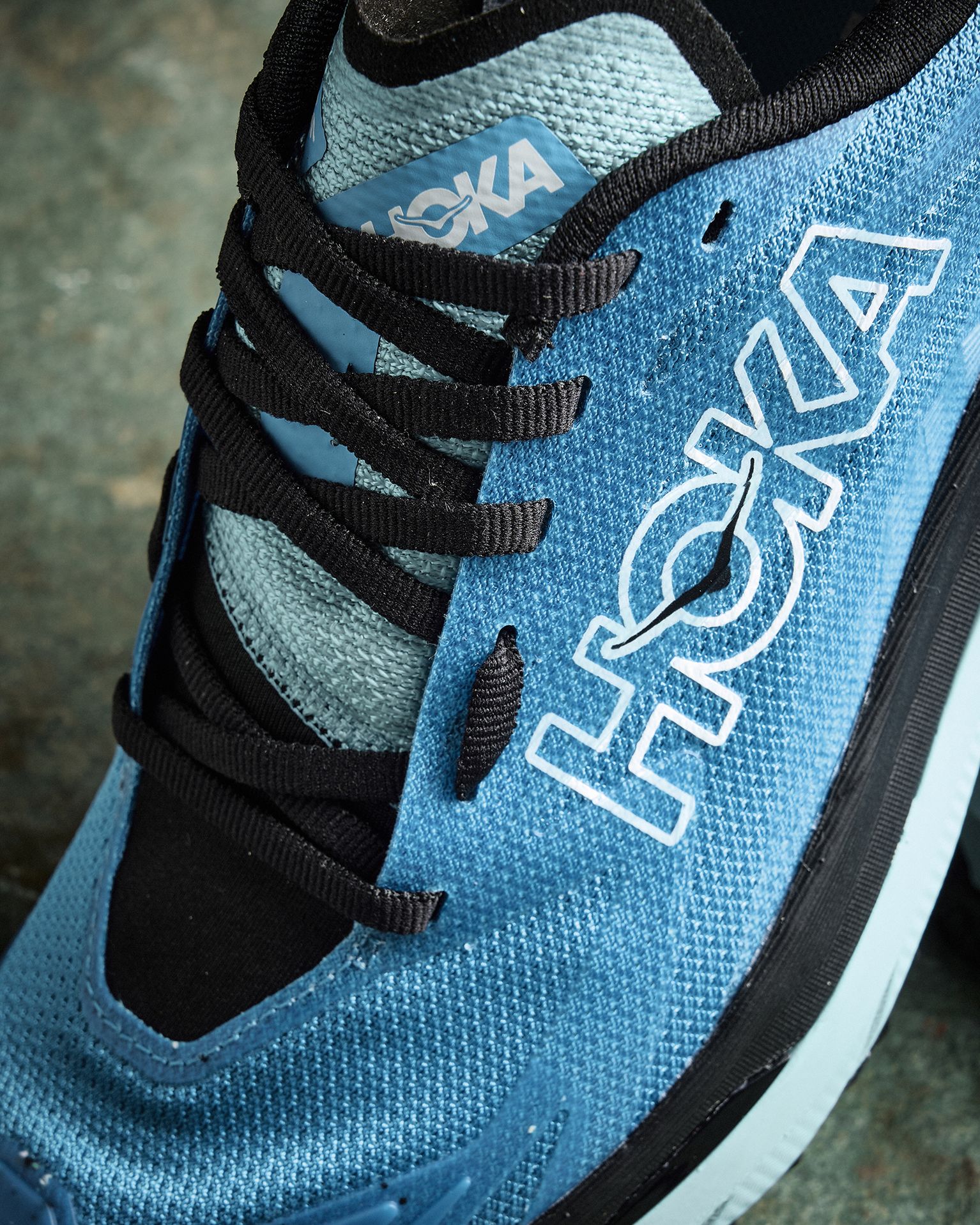 Close-up of a blue Hoka running shoe with black laces on a textured surface. The brand name is prominently displayed on the side.