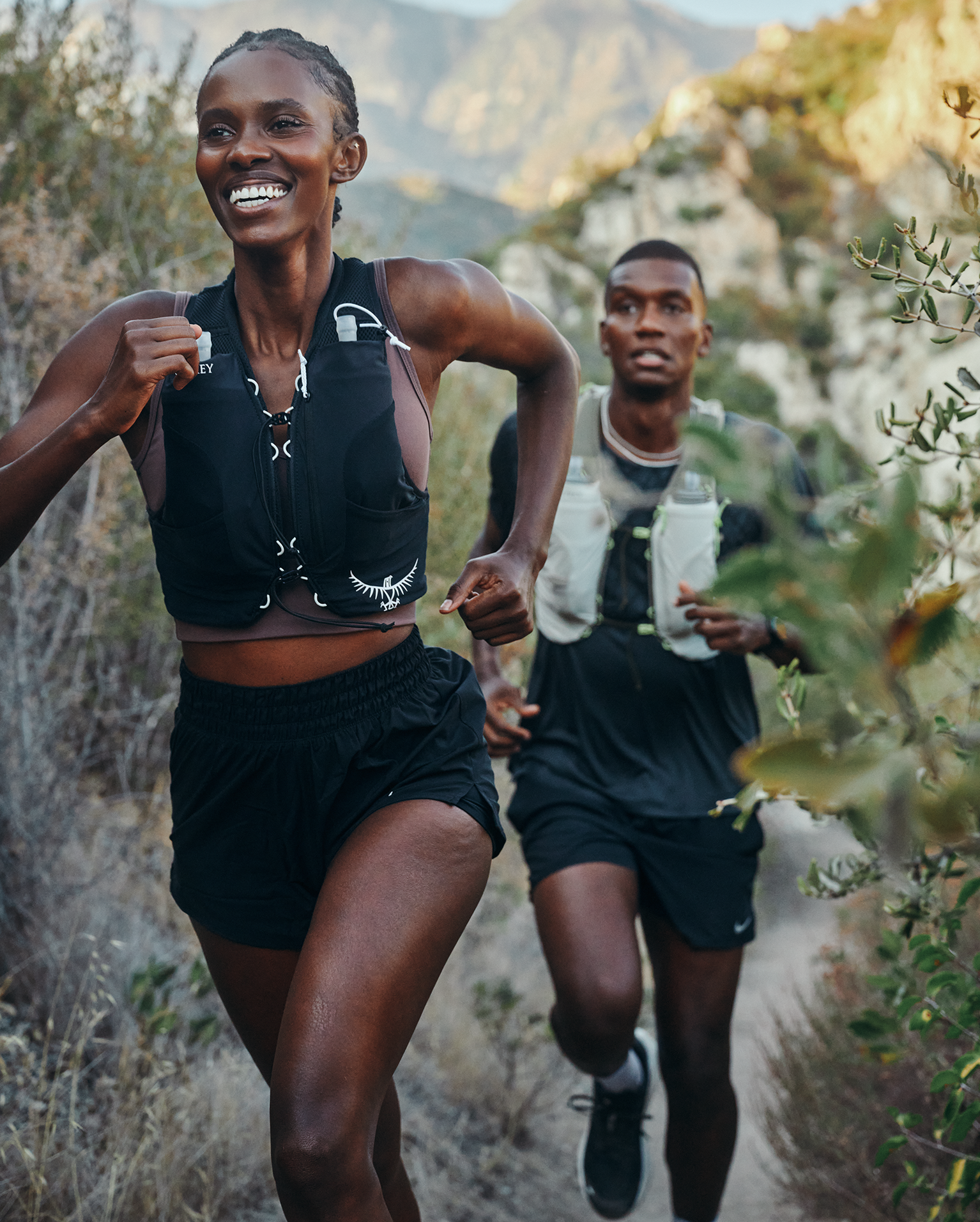Two people trail running energetically on a scenic mountain path, wearing running gear and hydration vests, with mountains in the background.