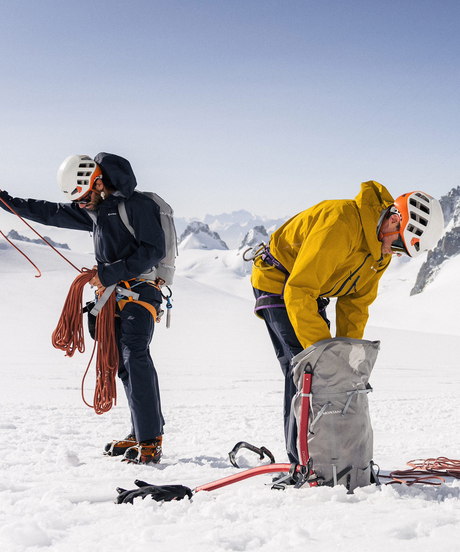Two climbers in helmets and gear prepare ropes on a snowy mountain. One stands holding a rope, the other crouches near a backpack.