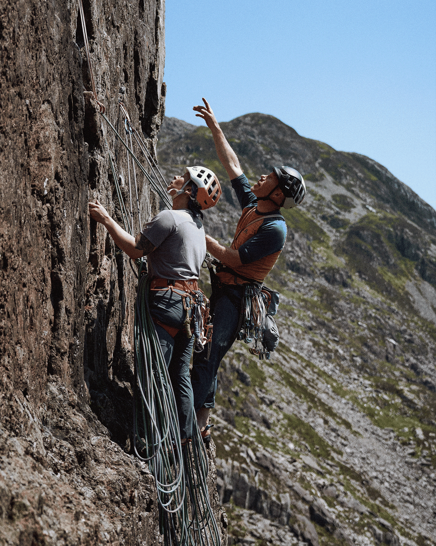 Two climbers, wearing helmets and harnesses, scale a rocky mountain face on a clear day, surrounded by rugged terrain.