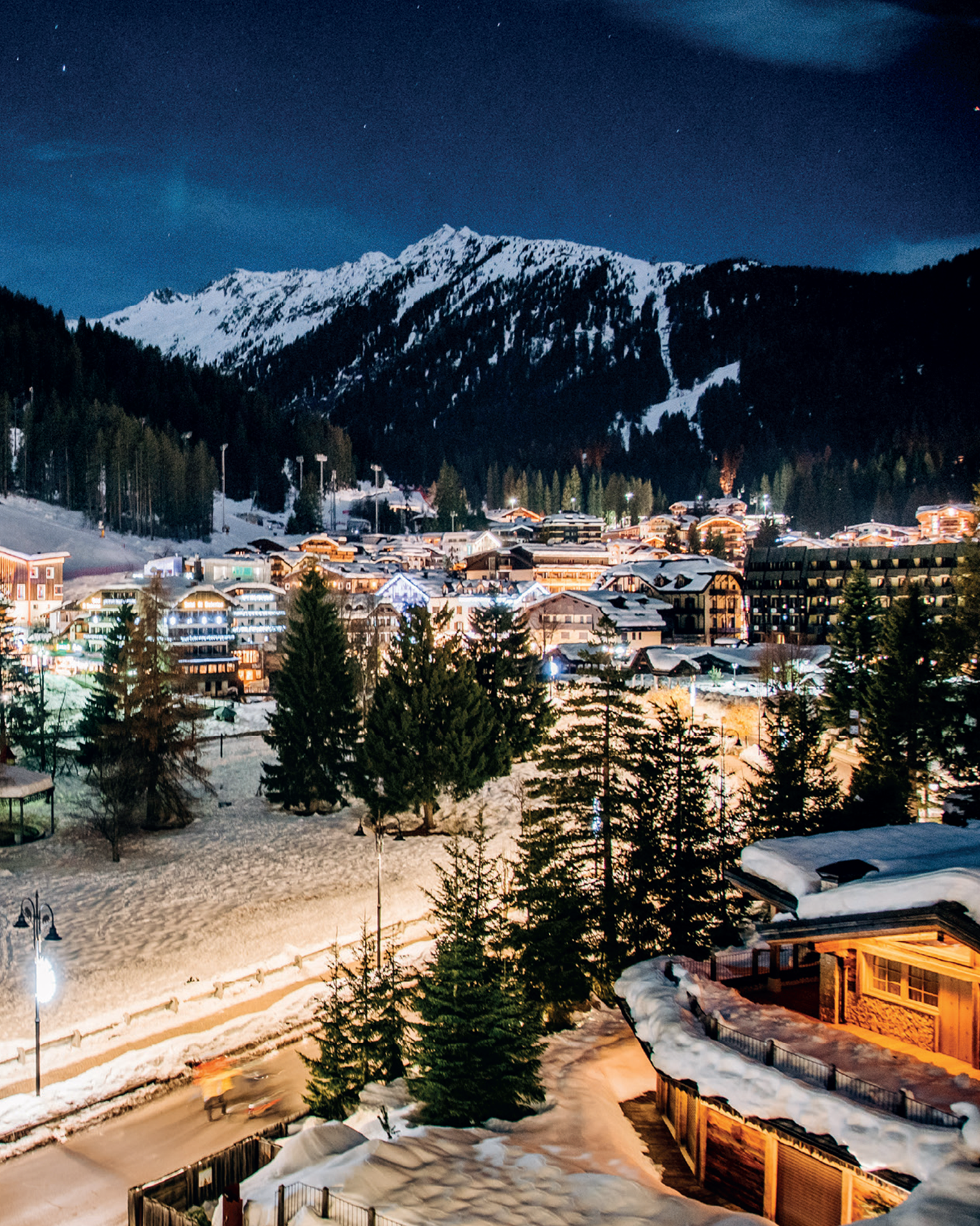 A picturesque alpine village illuminated at night, nestled below snow-covered mountains with trees and buildings in a serene landscape.