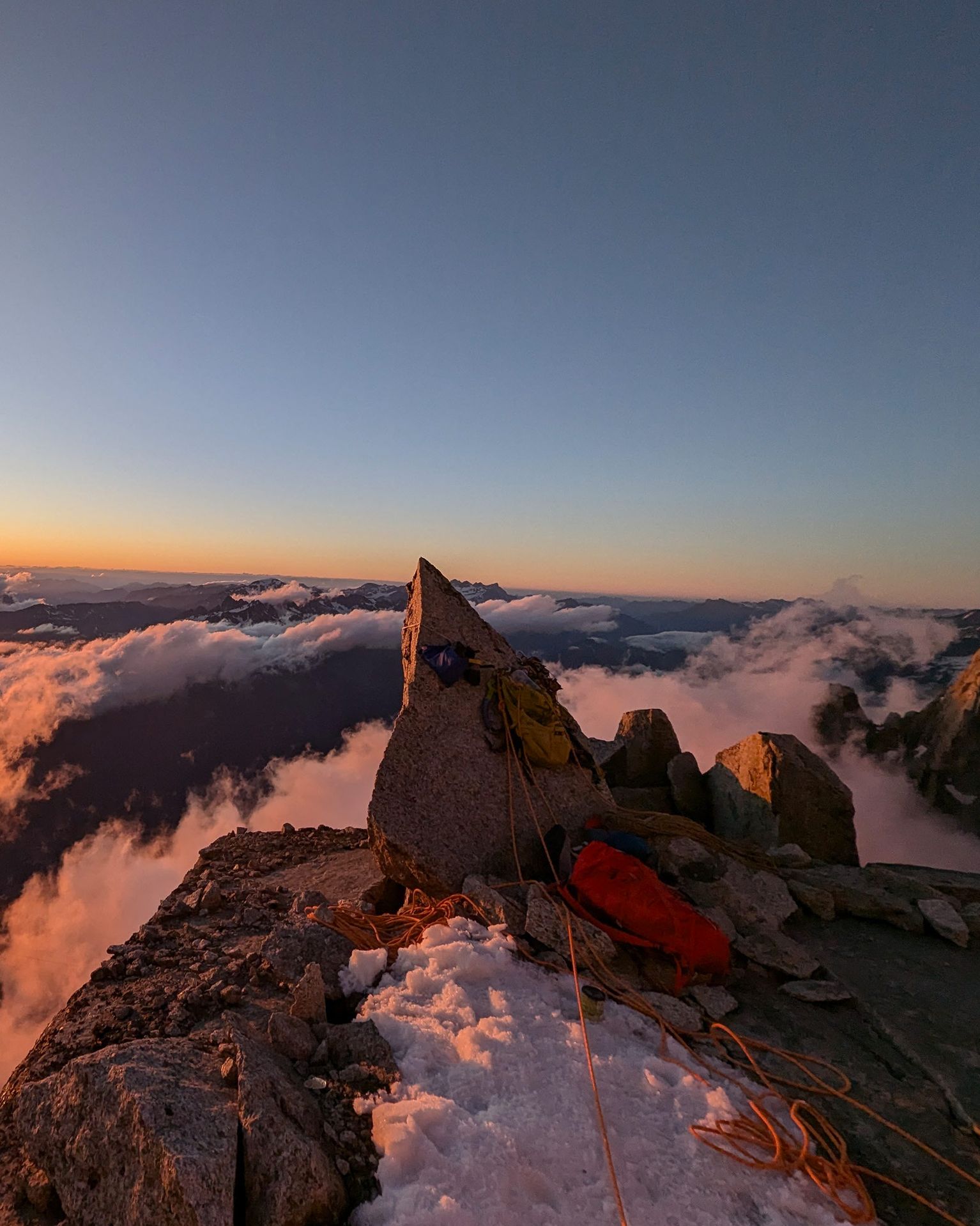 Sunrise view from a mountain peak with climbing gear, snow, and clouds below, casting a warm glow over the rugged landscape.