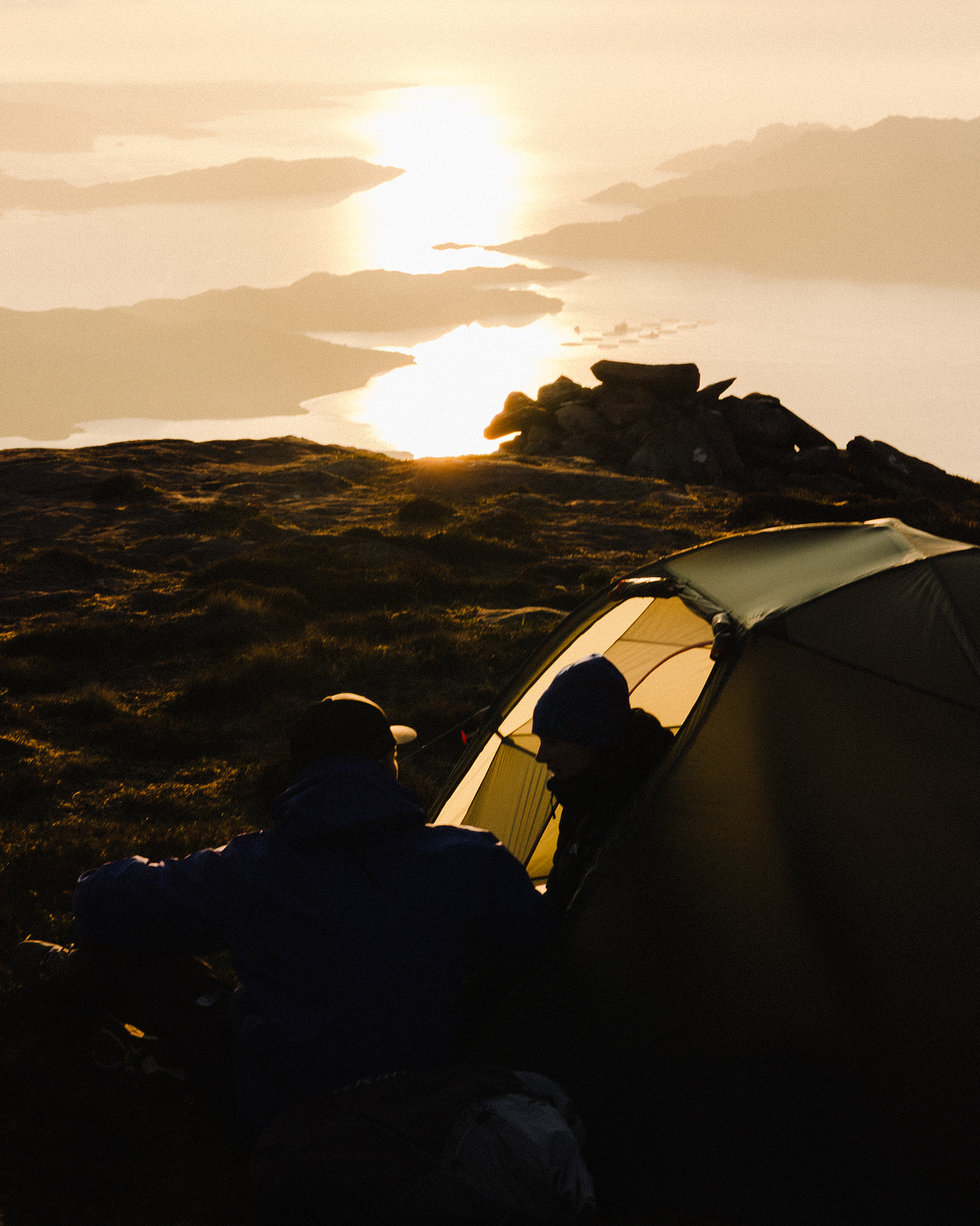 Campers sitting by a tent on a mountain, overlooking a tranquil sunset and still waters below.