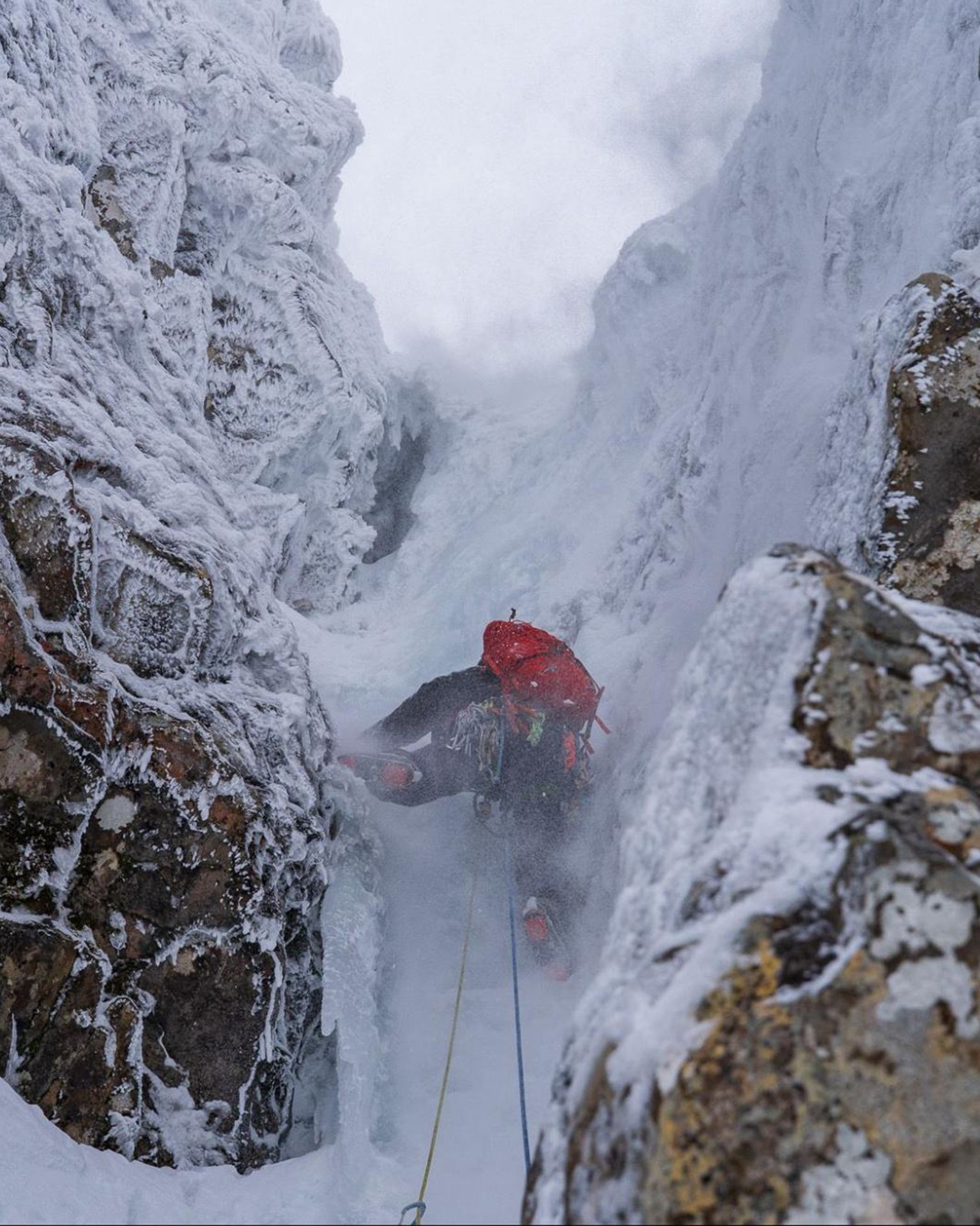 Climber ascending a narrow, snow-covered gully between rocky cliffs in winter conditions, with a red backpack and ropes.