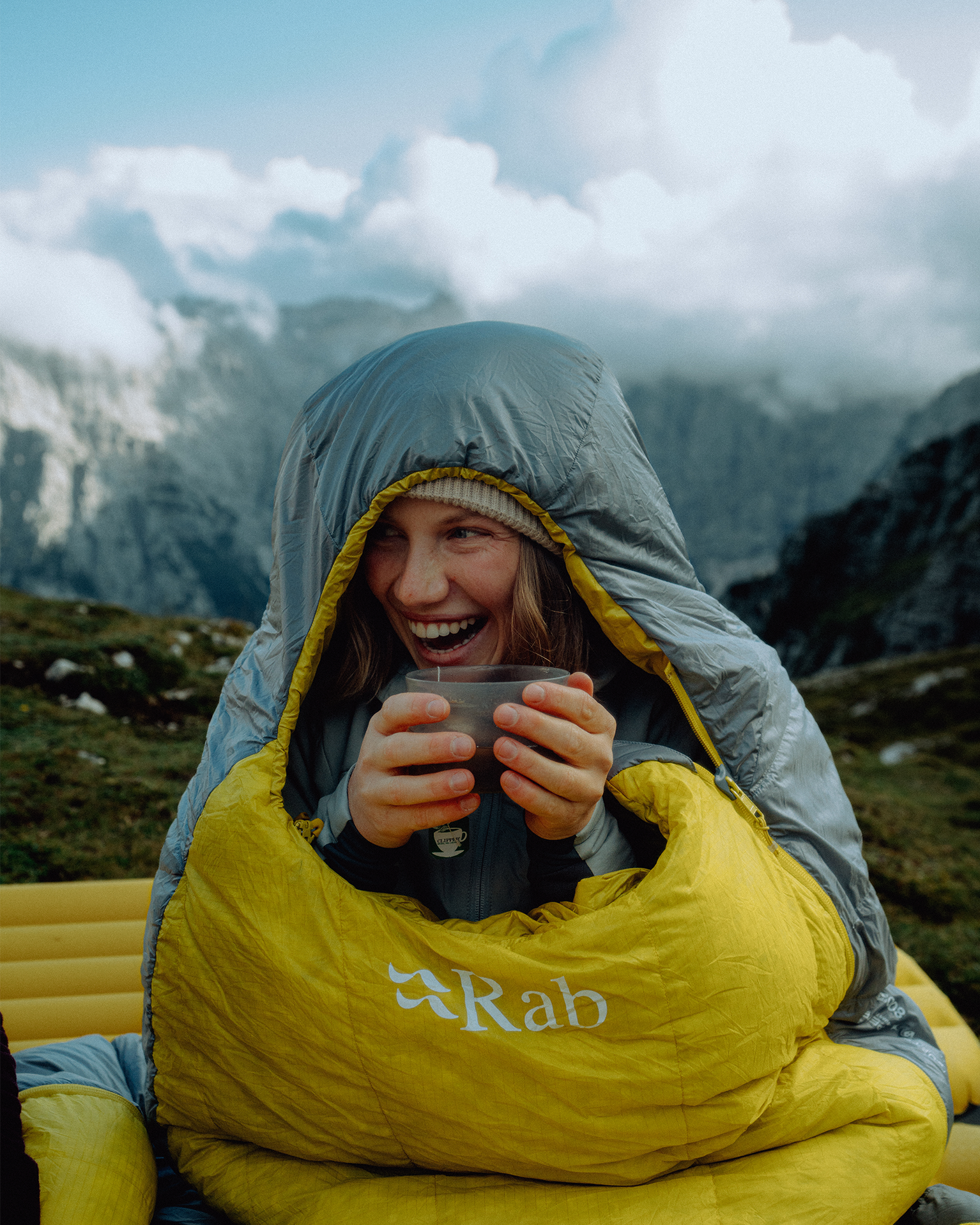 Woman wrapped in a yellow sleeping bag, smiling, holding a cup, sitting outdoors with mountains and clouds in the background.