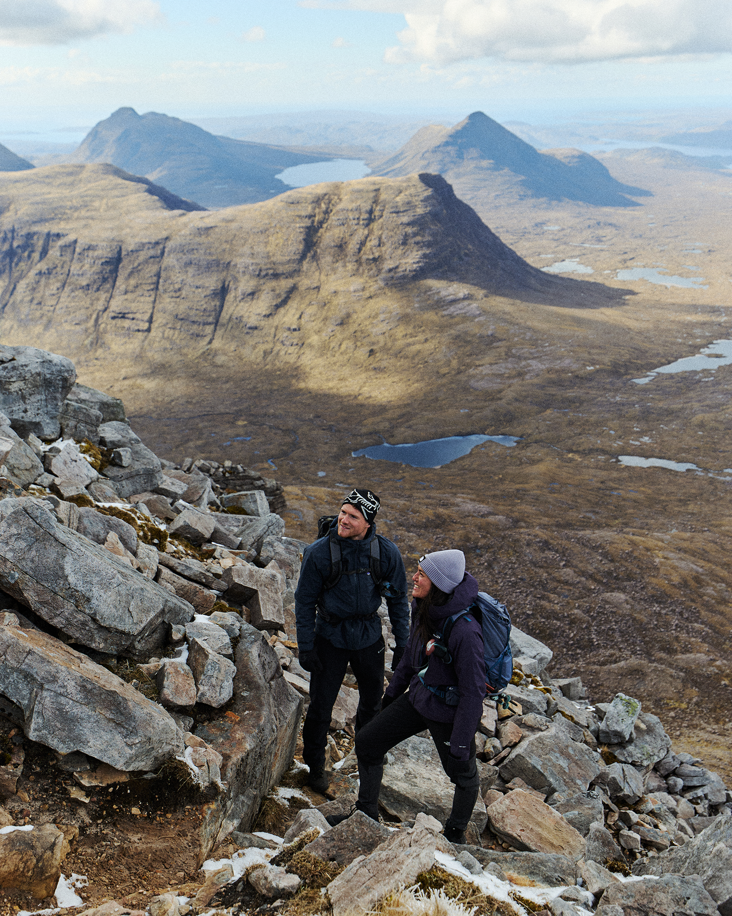 Two hikers in mountain gear traverse rocky terrain, surrounded by vast, rugged landscape and distant peaks under a blue sky with clouds.