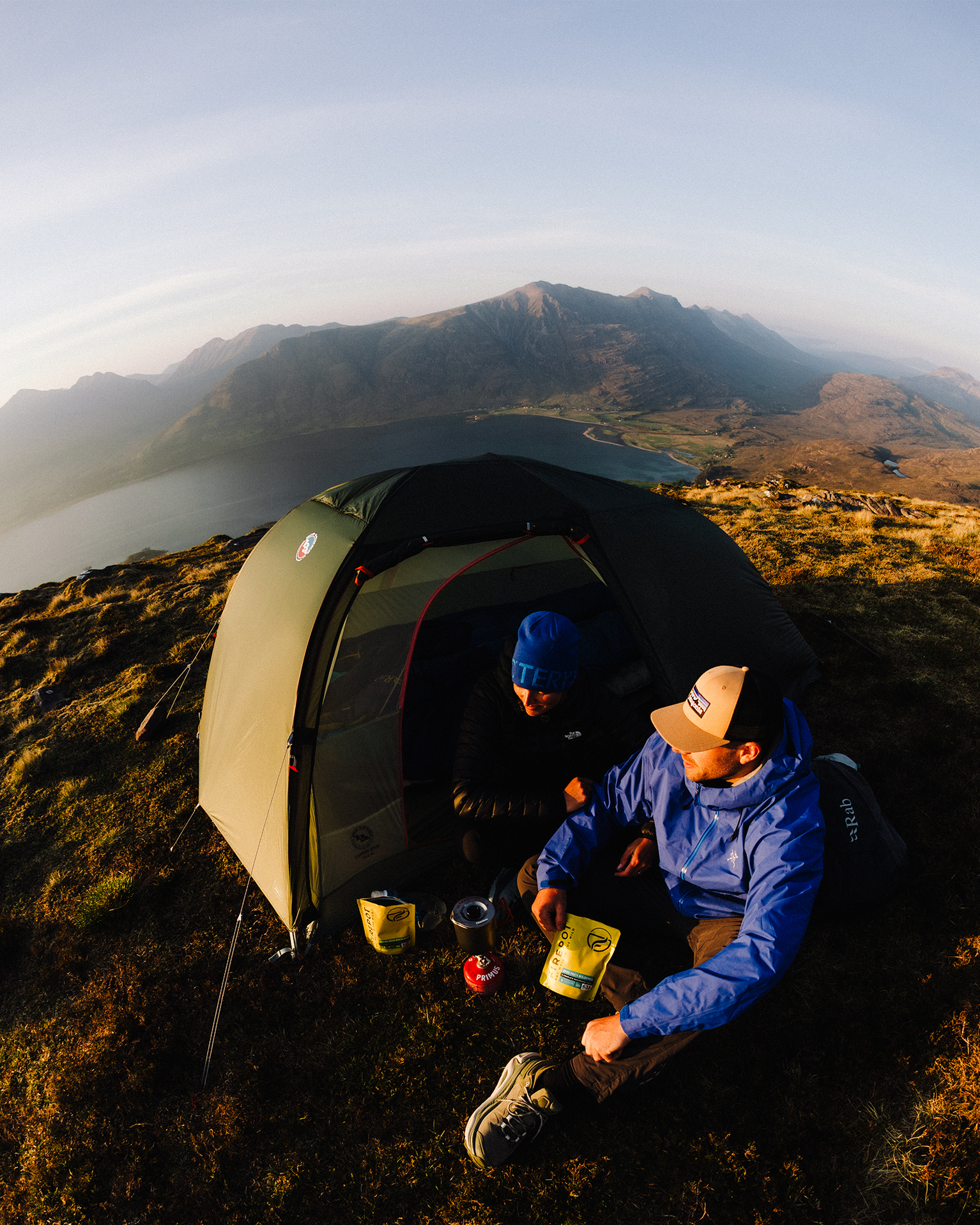 Two campers in jackets sit by a green tent on a mountain, enjoying a meal with scenic views of a lake and mountains in the background.
