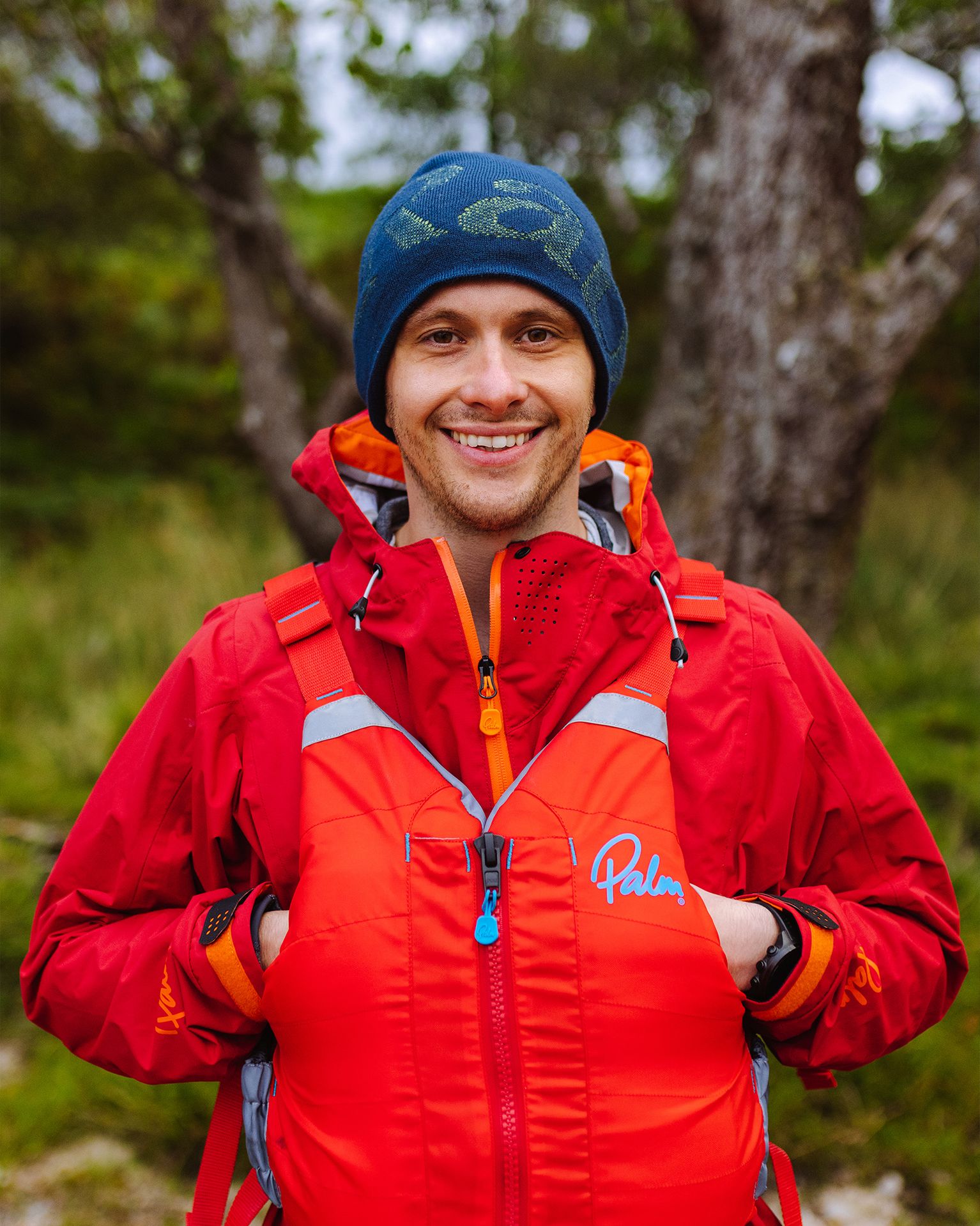 Smiling person in a red jacket and beanie stands outdoors, hands in pockets, with trees and greenery in the background.