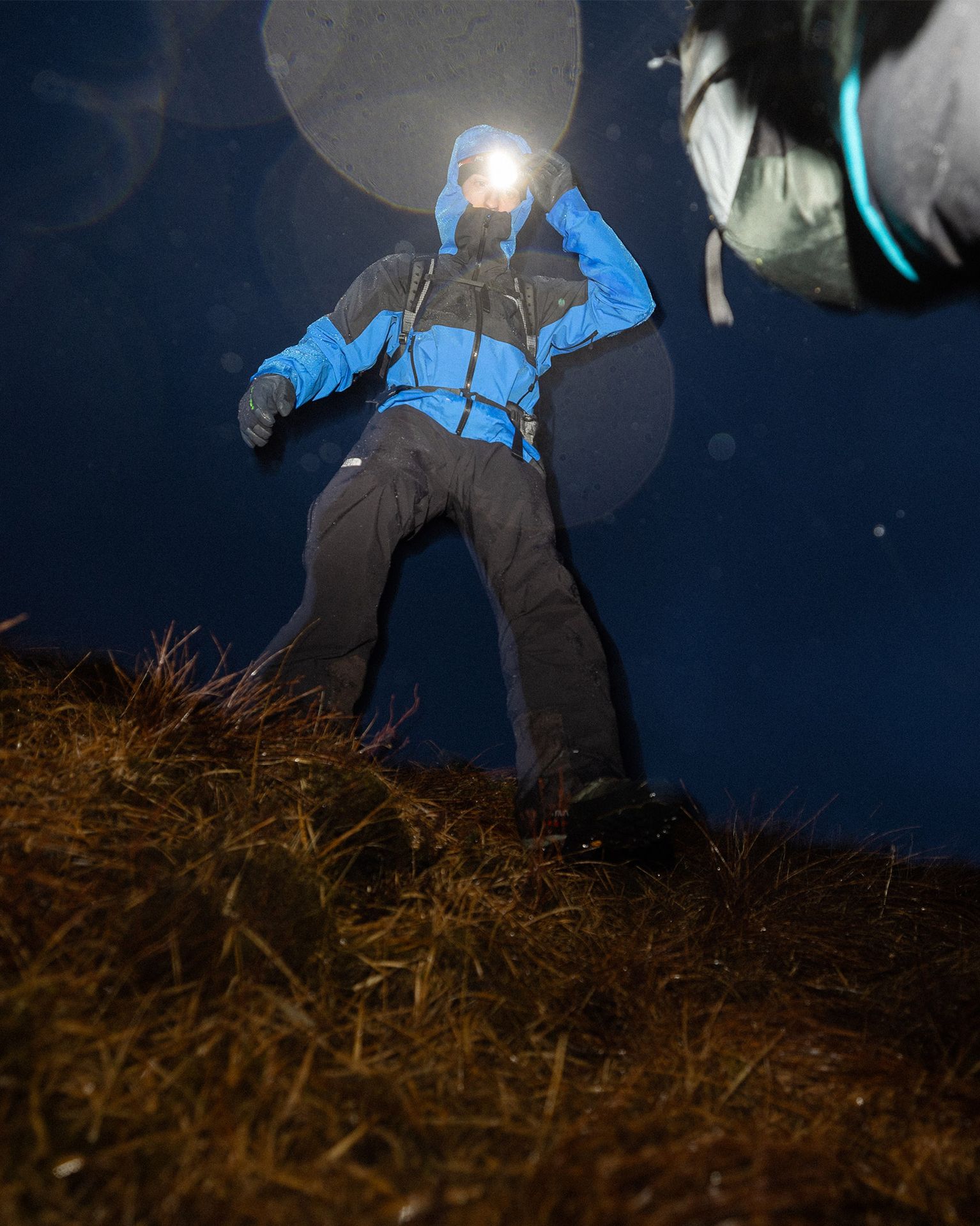Person in blue outdoor gear shines a head torch at the camera, standing on a grassy slope at night.