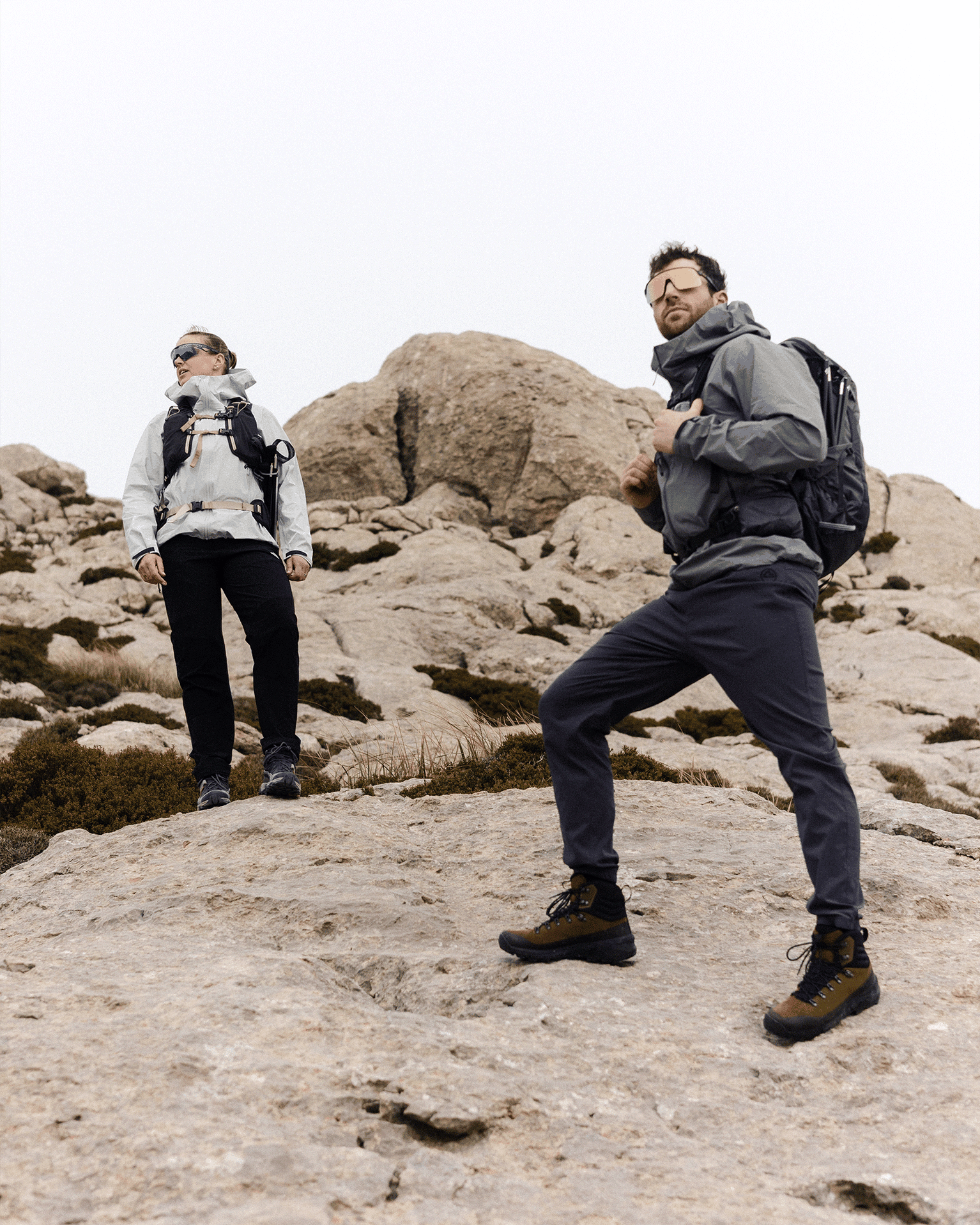 Two hikers in outdoor gear stand on rocky terrain, looking into the distance under an overcast sky.