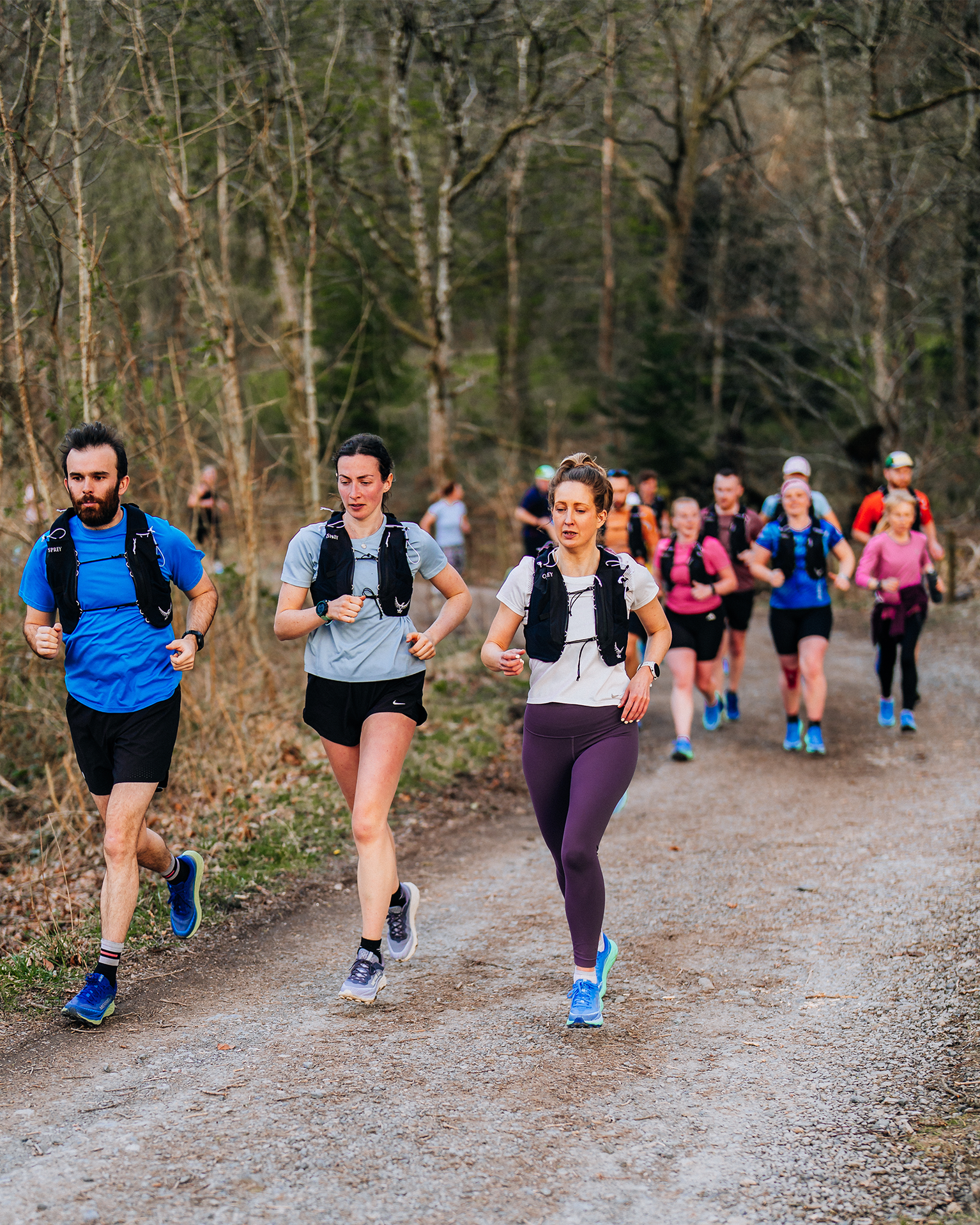 Group of trail runners jogging through a forest path, wearing sporting attire and backpacks, surrounded by trees.