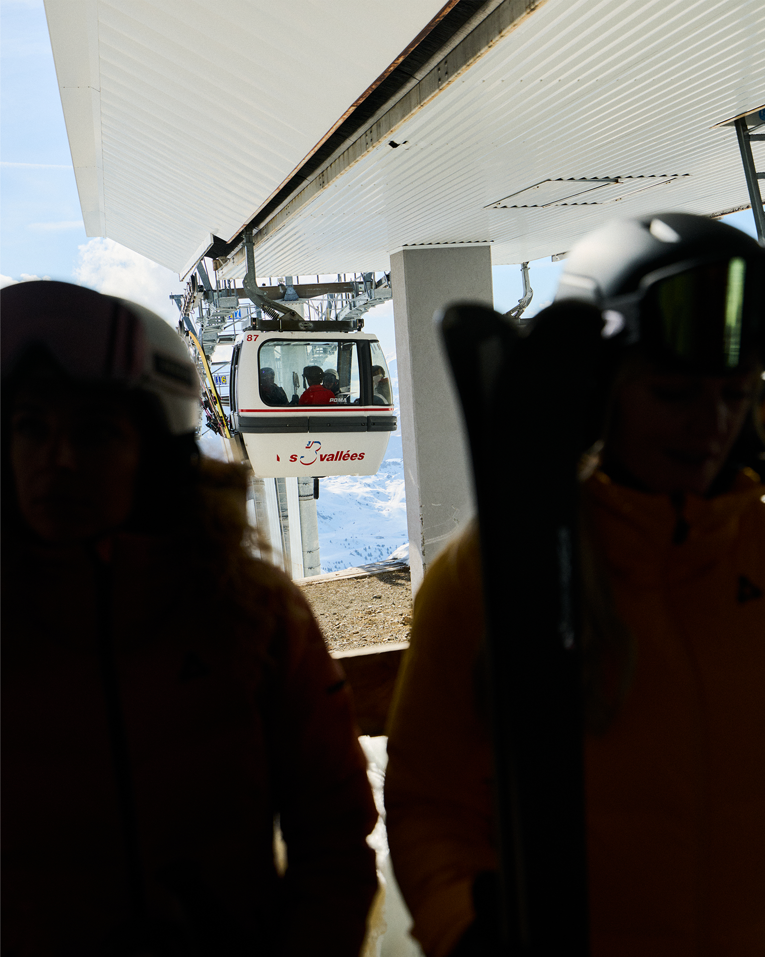 Two skiers in helmets near a gondola station, cable car moving in the background with snowy mountain scenery.