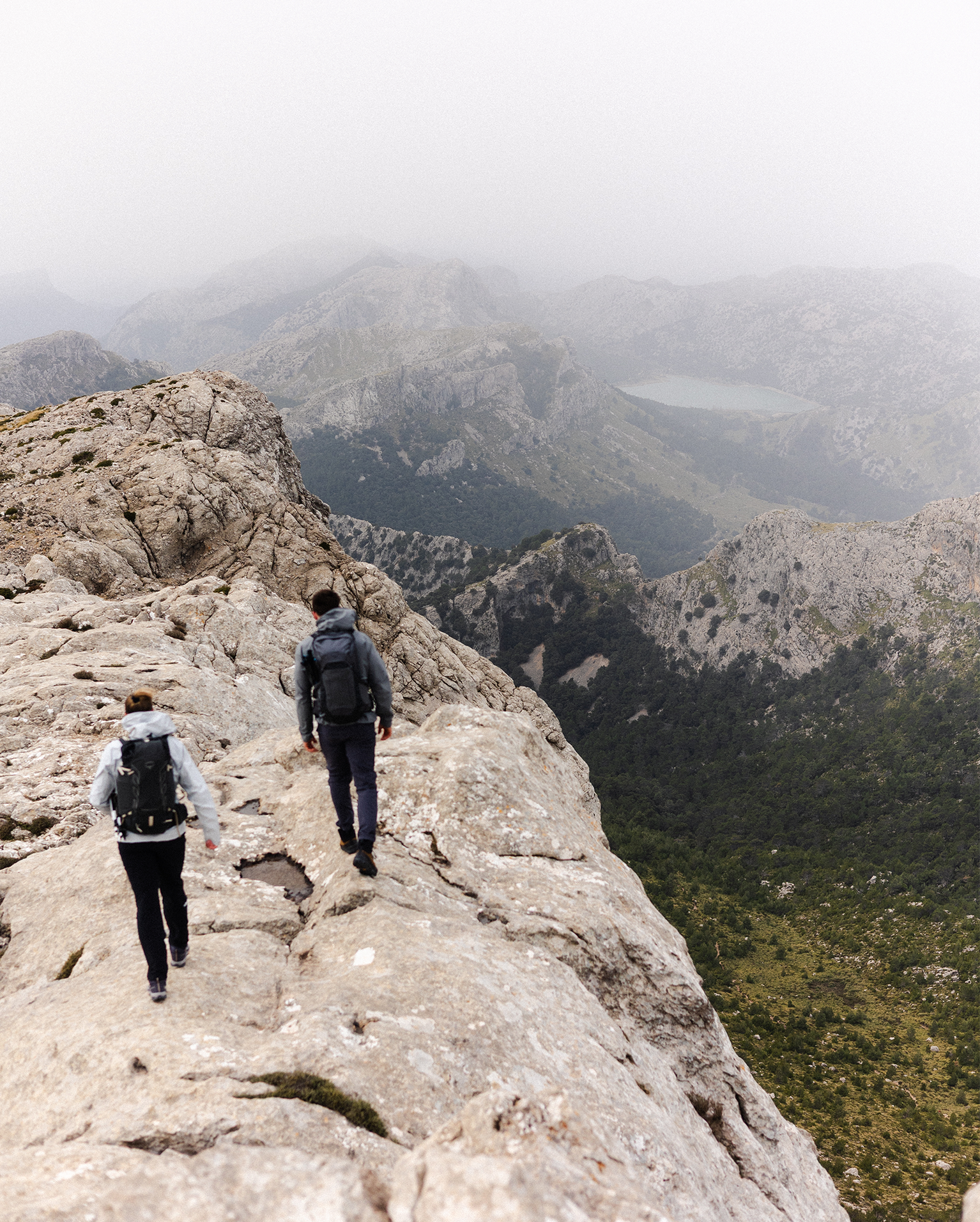 Two hikers with backpacks walking along a rocky mountain ridge, overlooking a scenic valley with misty skies.