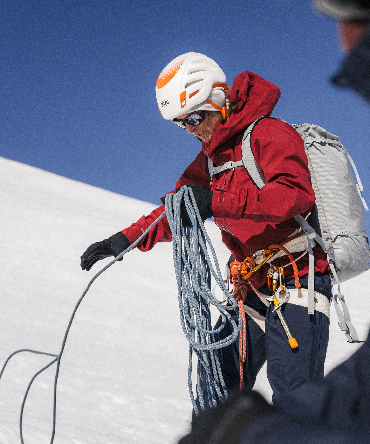 A climber in a red jacket and helmet handles a coiled rope on a snowy mountain under a clear blue sky.