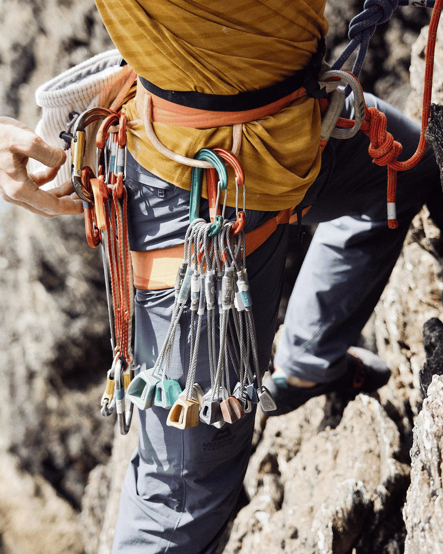 Climber wearing a harness with colourful carabiners and gear attached, scaling a rocky surface.