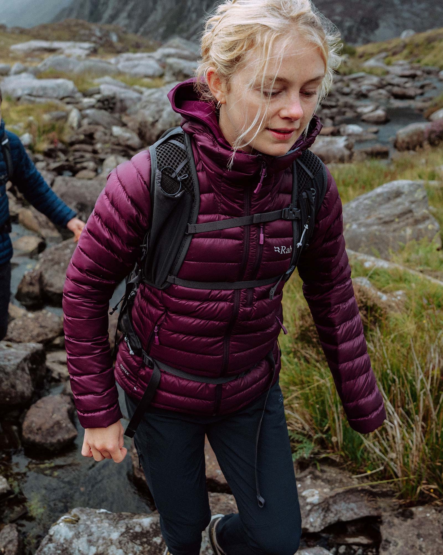 Woman hiking over rocky terrain in a maroon puffer jacket and backpack, surrounded by grass and stones.