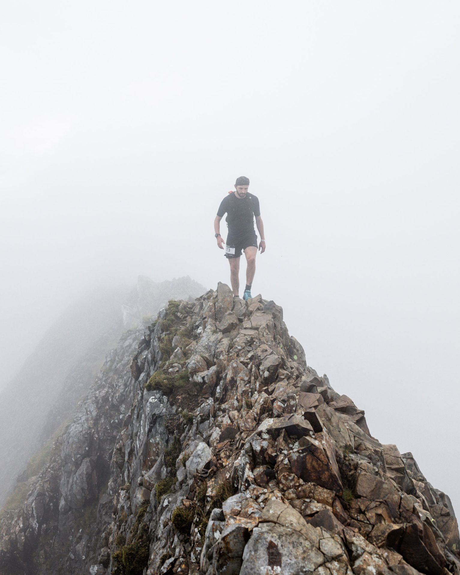 A person walks along a narrow, rocky mountain ridge enveloped in fog, wearing a black shirt and shorts, with a determined expression.