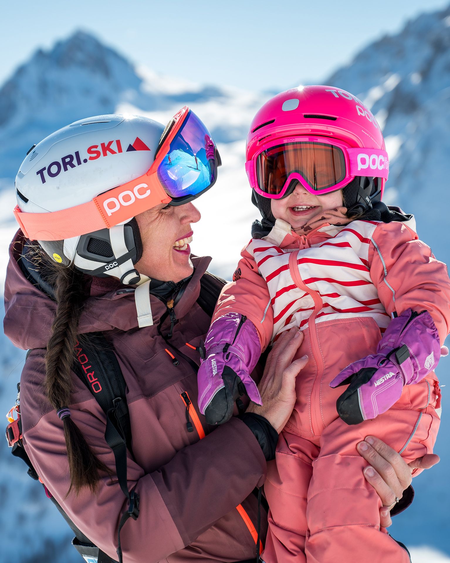A woman in ski gear joyfully holds a child, both in helmets and goggles, against a snowy mountain backdrop.