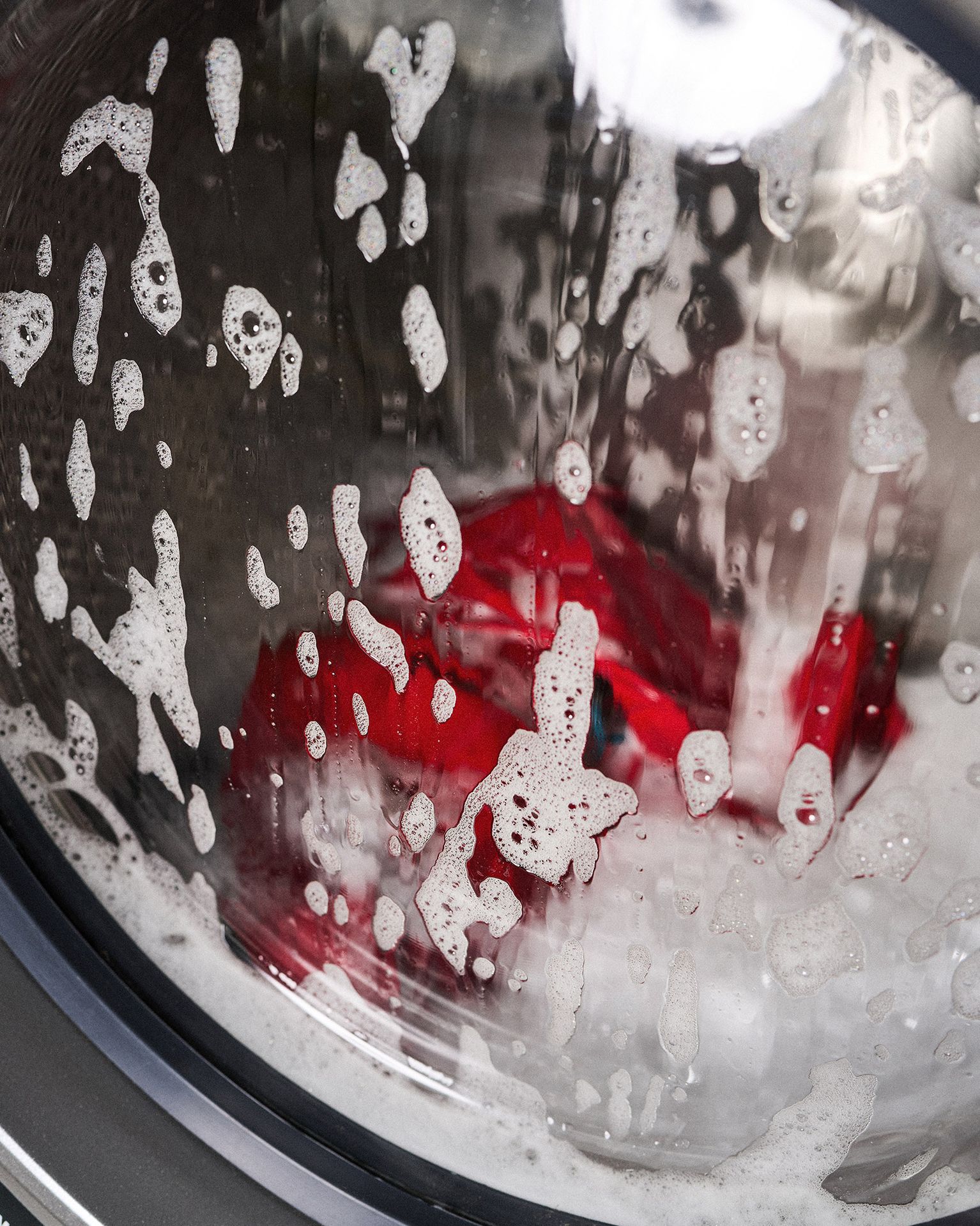 Washing machine door with soap suds and a red garment visible inside during a wash cycle.