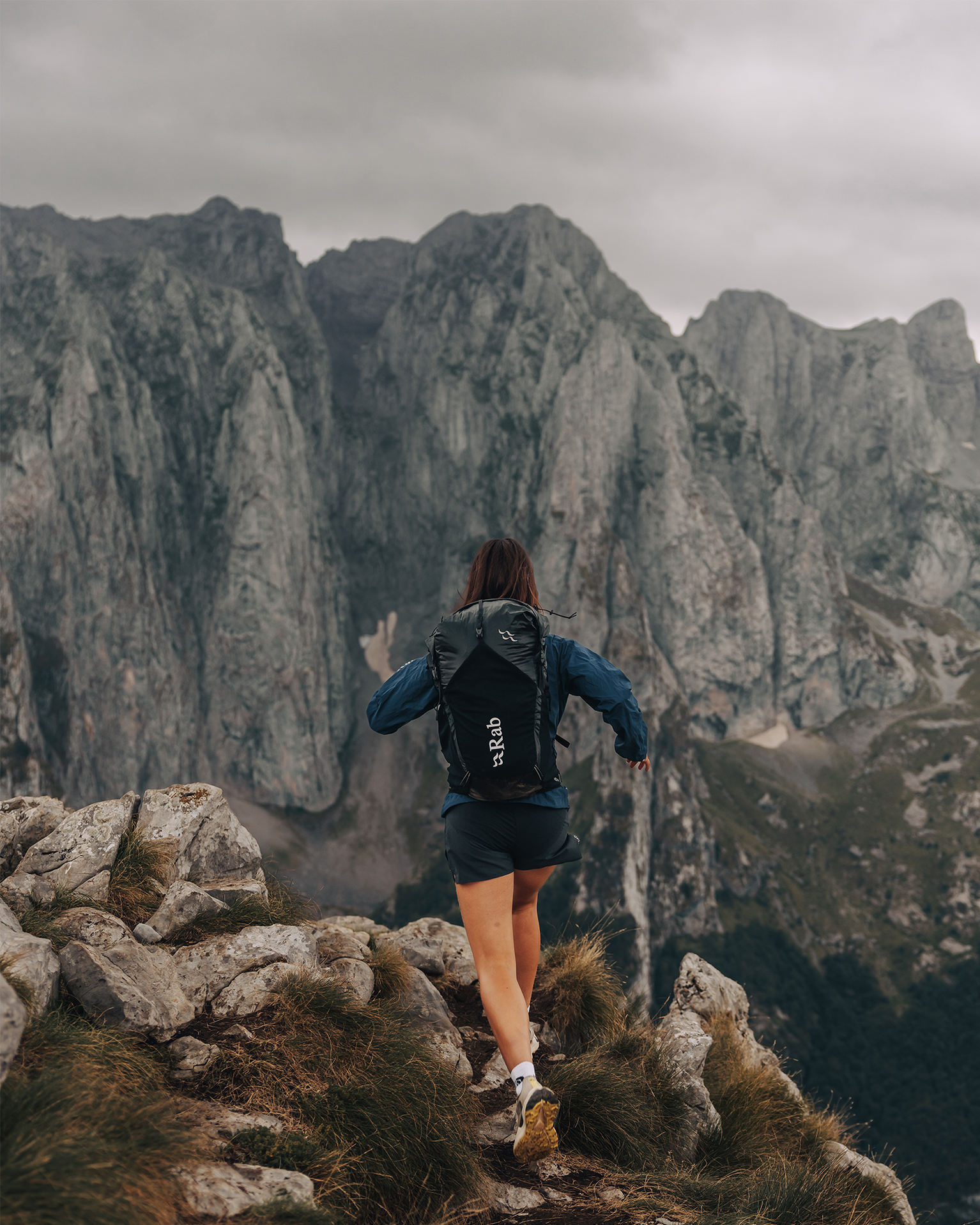 Person in mountain sports gear running on rugged terrain with towering rocky peaks in the background.