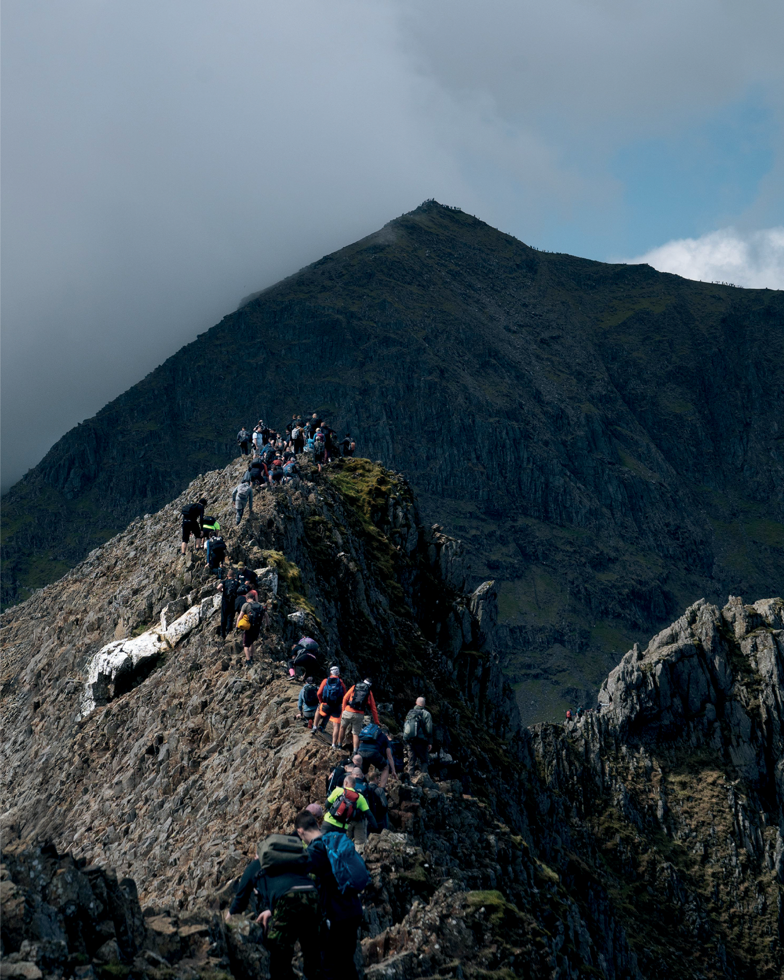 Hikers carefully navigate a narrow, rocky ridge leading to a mountain peak under a dramatic sky.