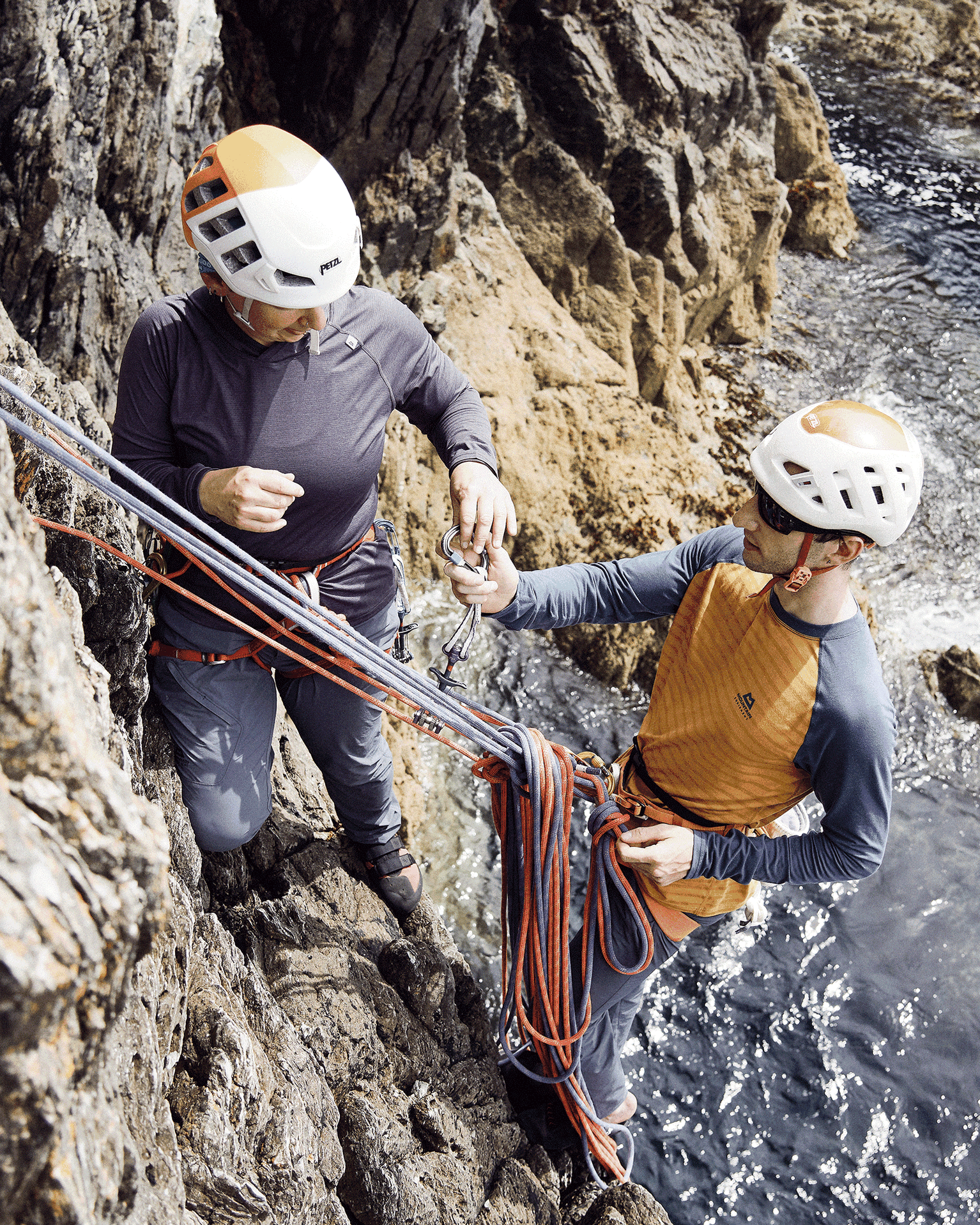 Two climbers in helmets and gear managing ropes on a rocky cliffside above the sea, demonstrating secure climbing techniques.